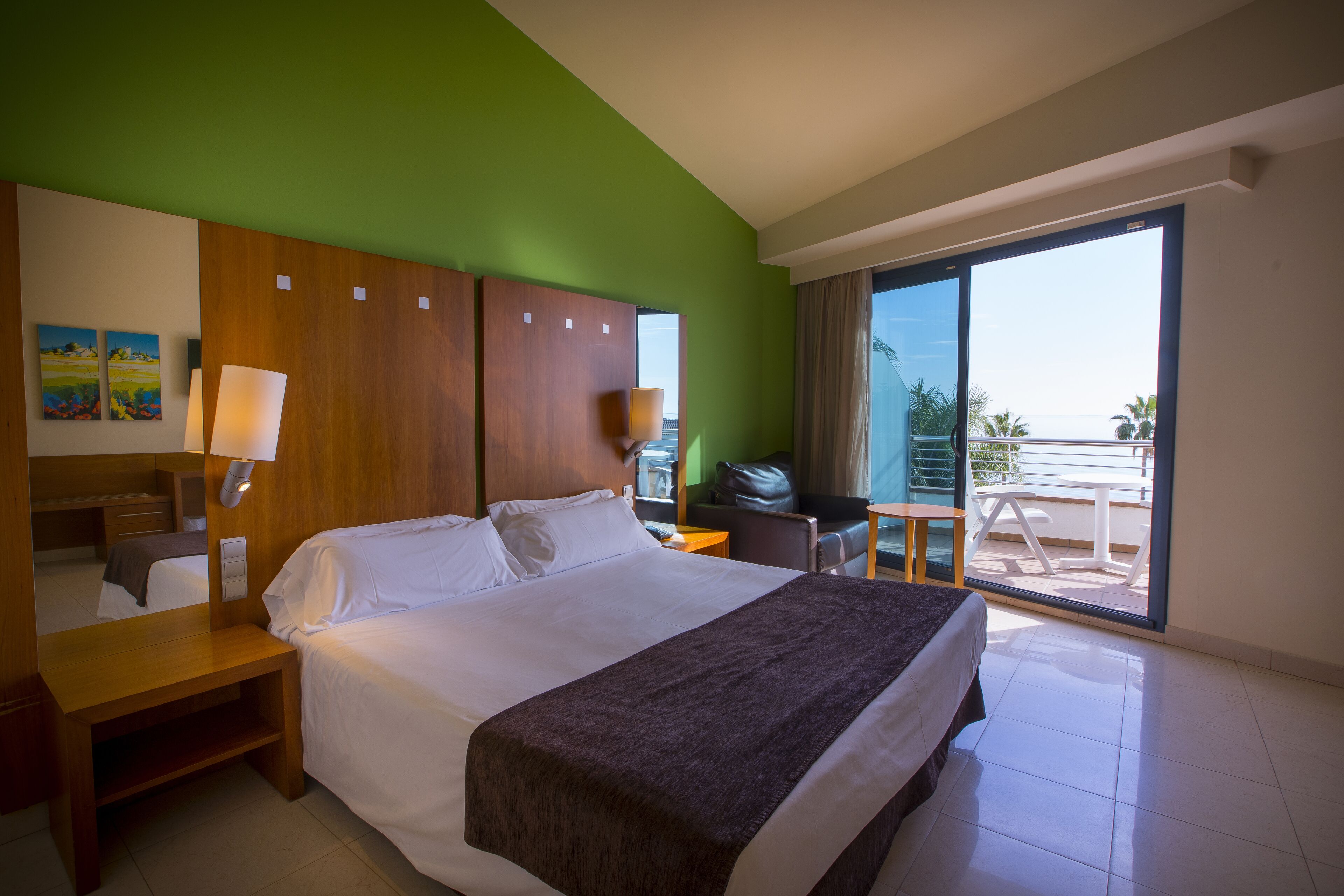 A hotel room with a large bed, a wooden headboard, a desk, a chair, a small table, and a balcony with a view of palm trees.