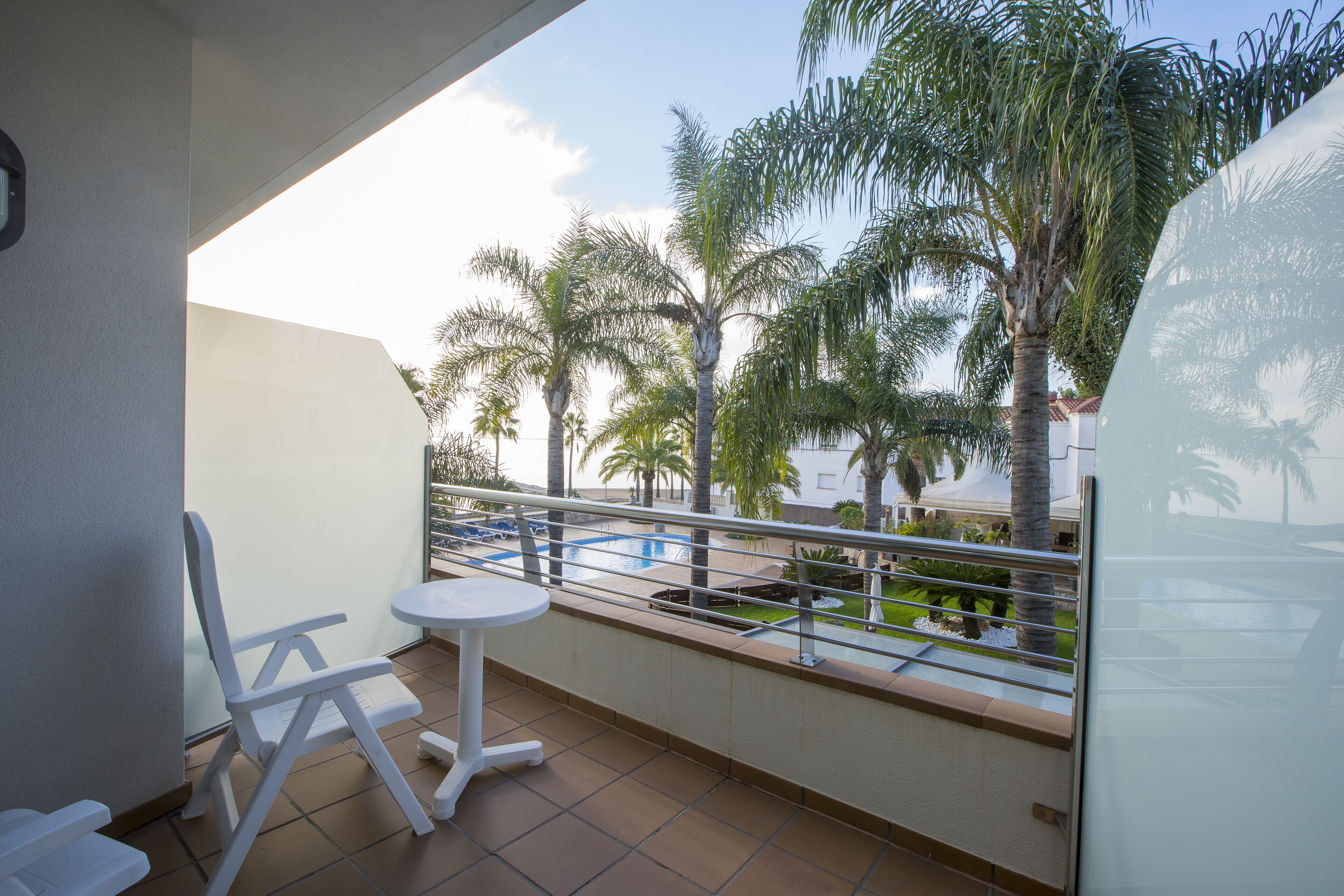A balcony with a white chair and table, overlooking a pool and palm trees.
