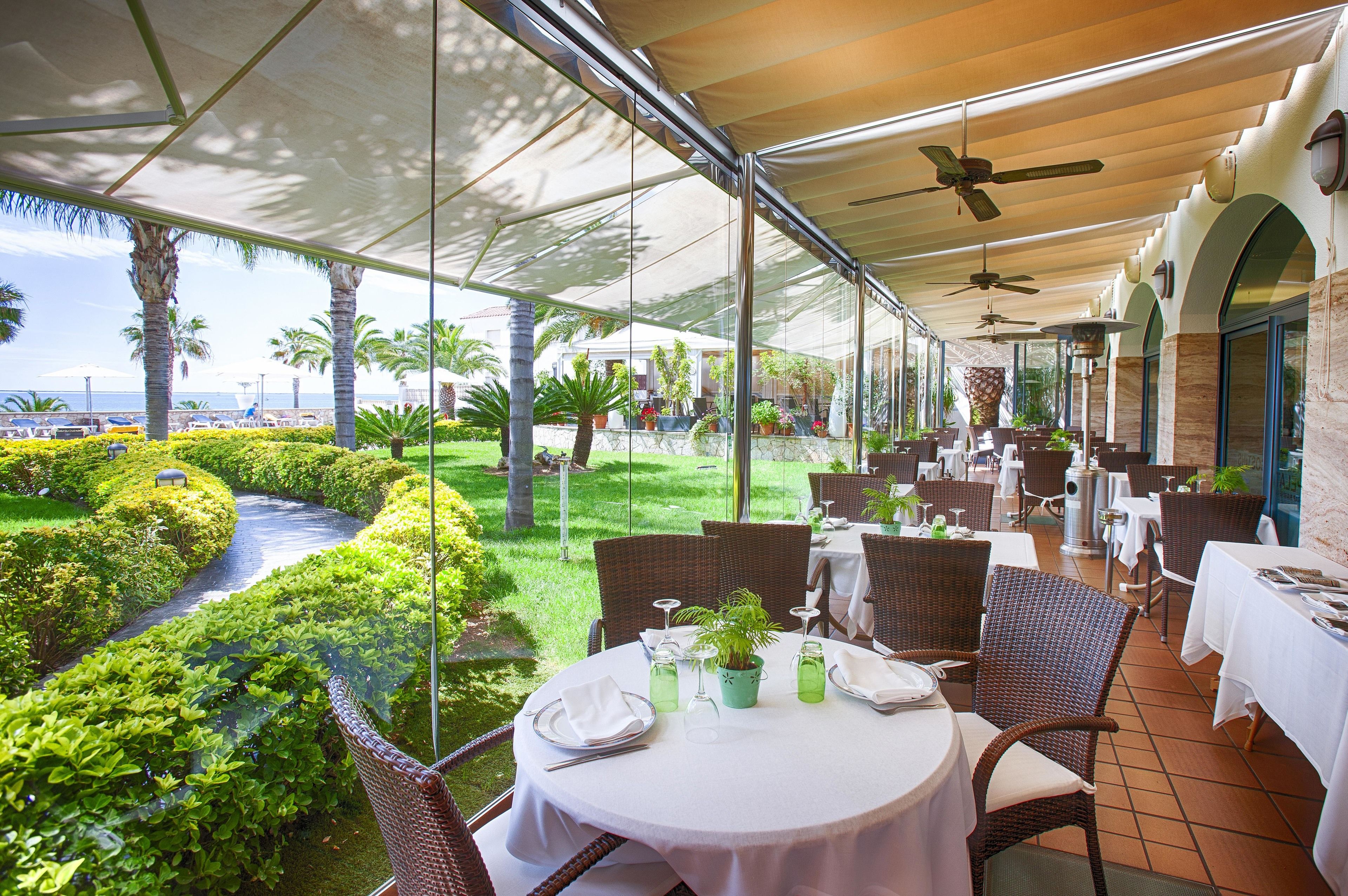 An outdoor dining area with tables and chairs, surrounded by greenery and a view of the sea.