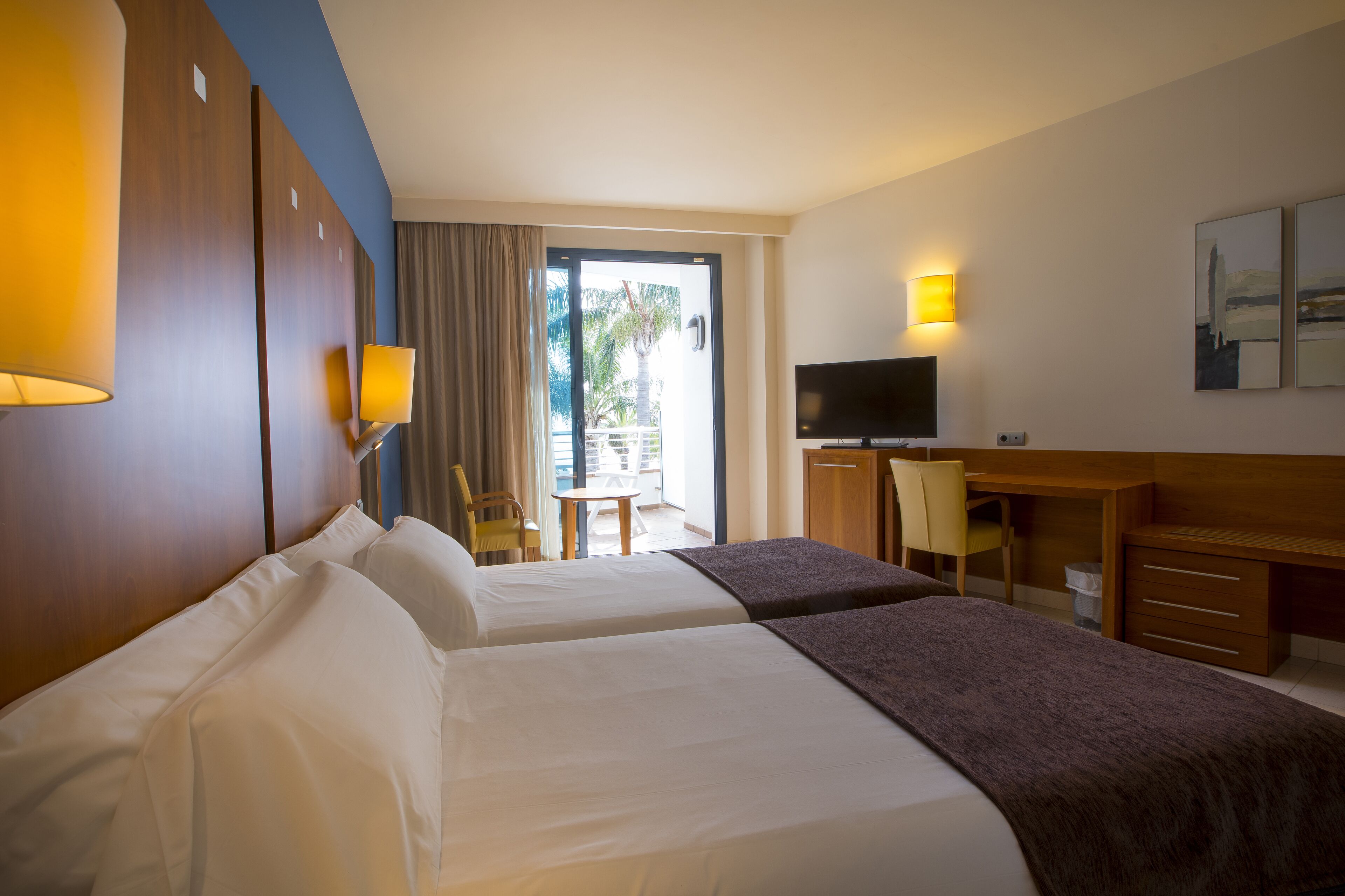 Une chambre d’hôtel avec deux lits, un bureau, une chaise, une télévision et un balcon donnant sur des palmiers.