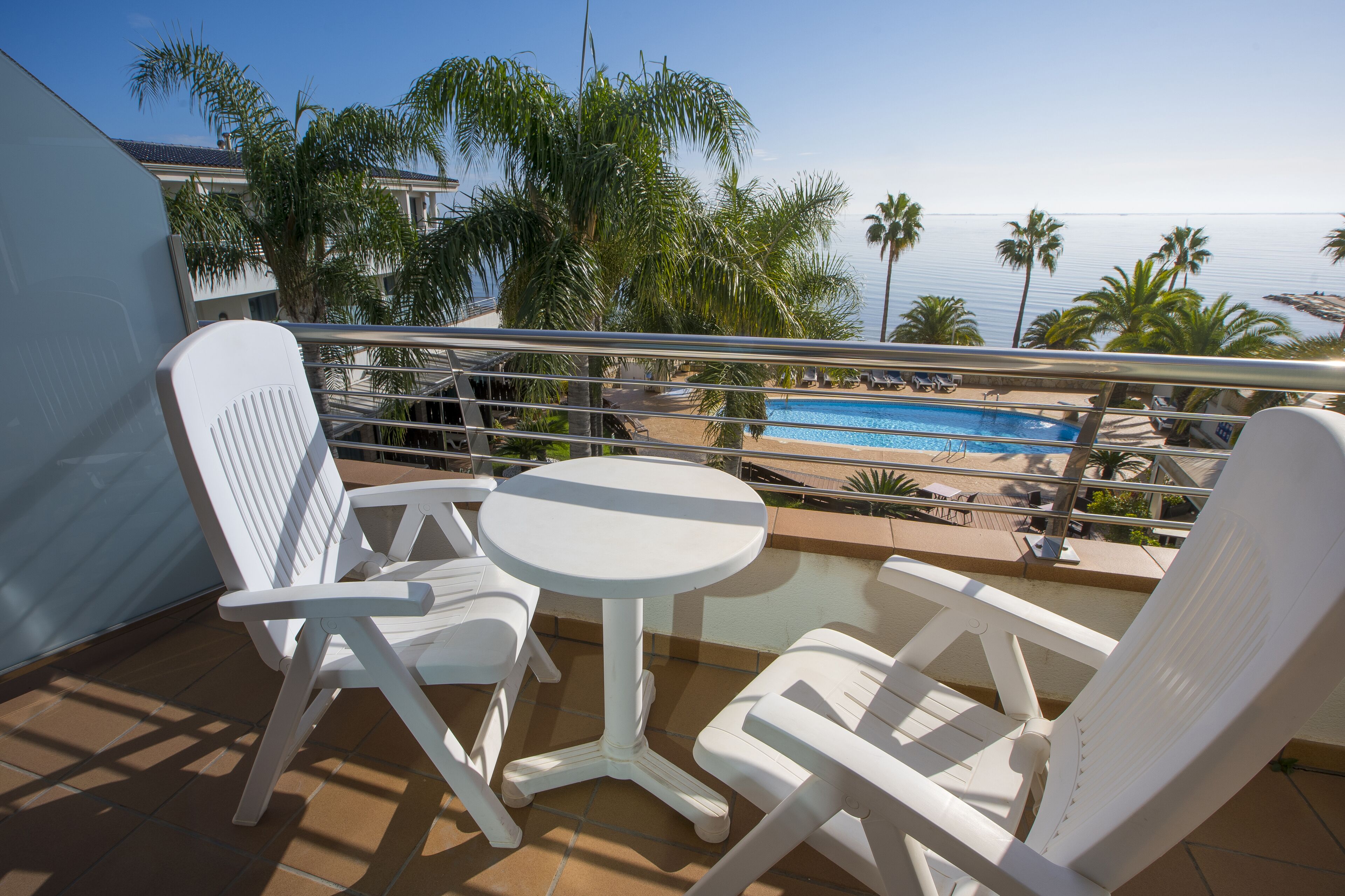 Un balcon avec des chaises blanches et une petite table, donnant sur une piscine et des palmiers.