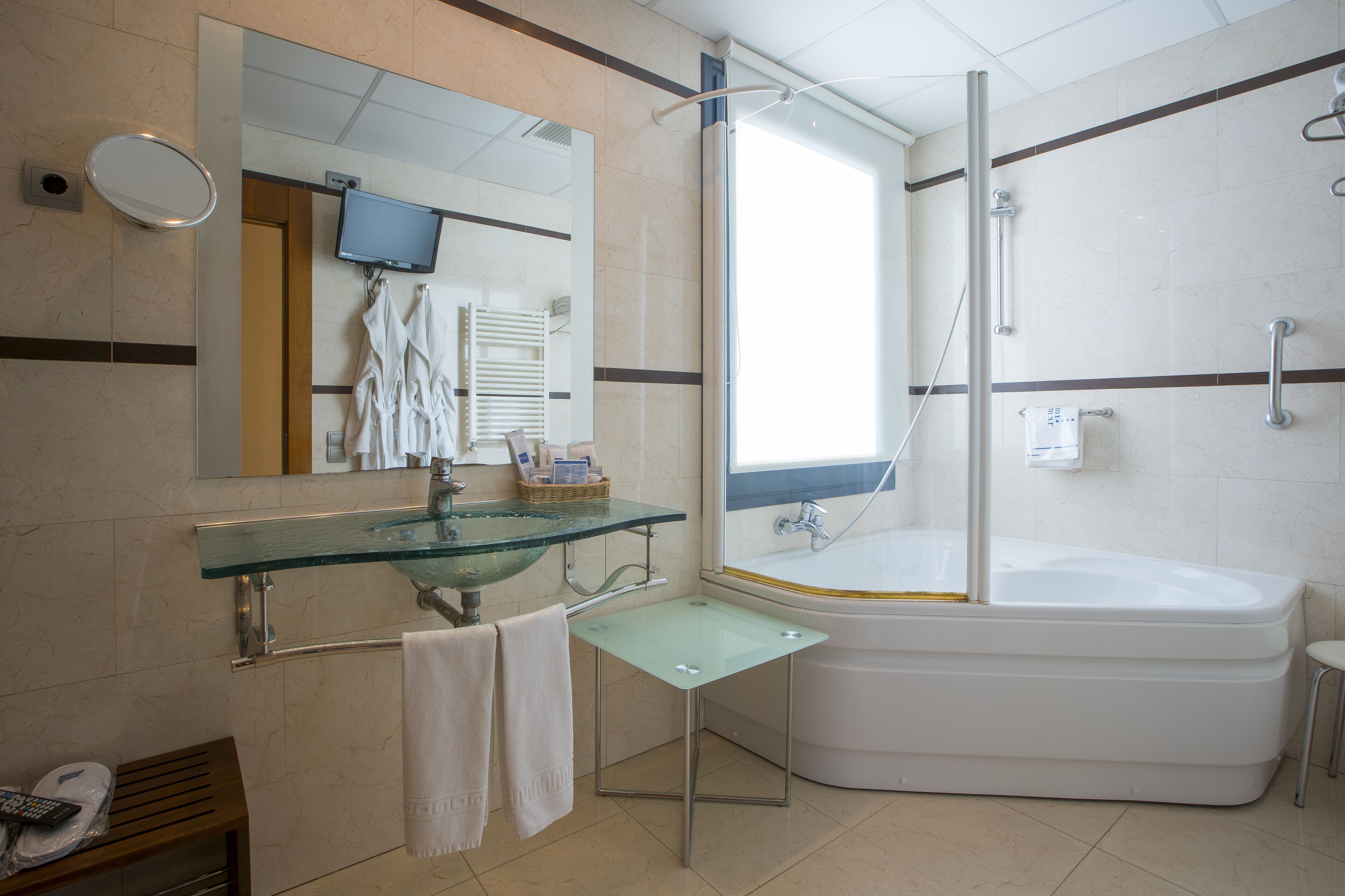 Un baño moderno con un lavamanos de vidrio, una bañera grande, un box de ducha y un espejo con televisión.