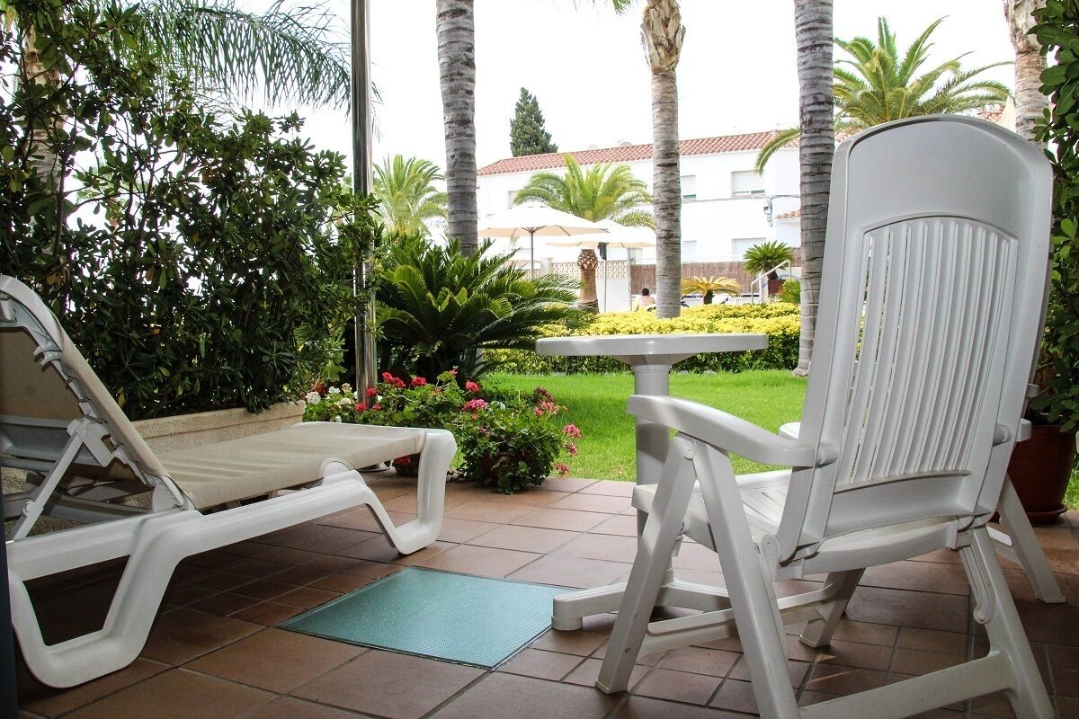 A patio area with a white lounge chair and a white rocking chair, surrounded by lush greenery and a well-maintained garden.