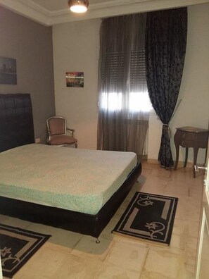A bedroom with a bed, a chair, a small table, and a window with curtains. - Benzineb Immo Menzeh (Ariana)