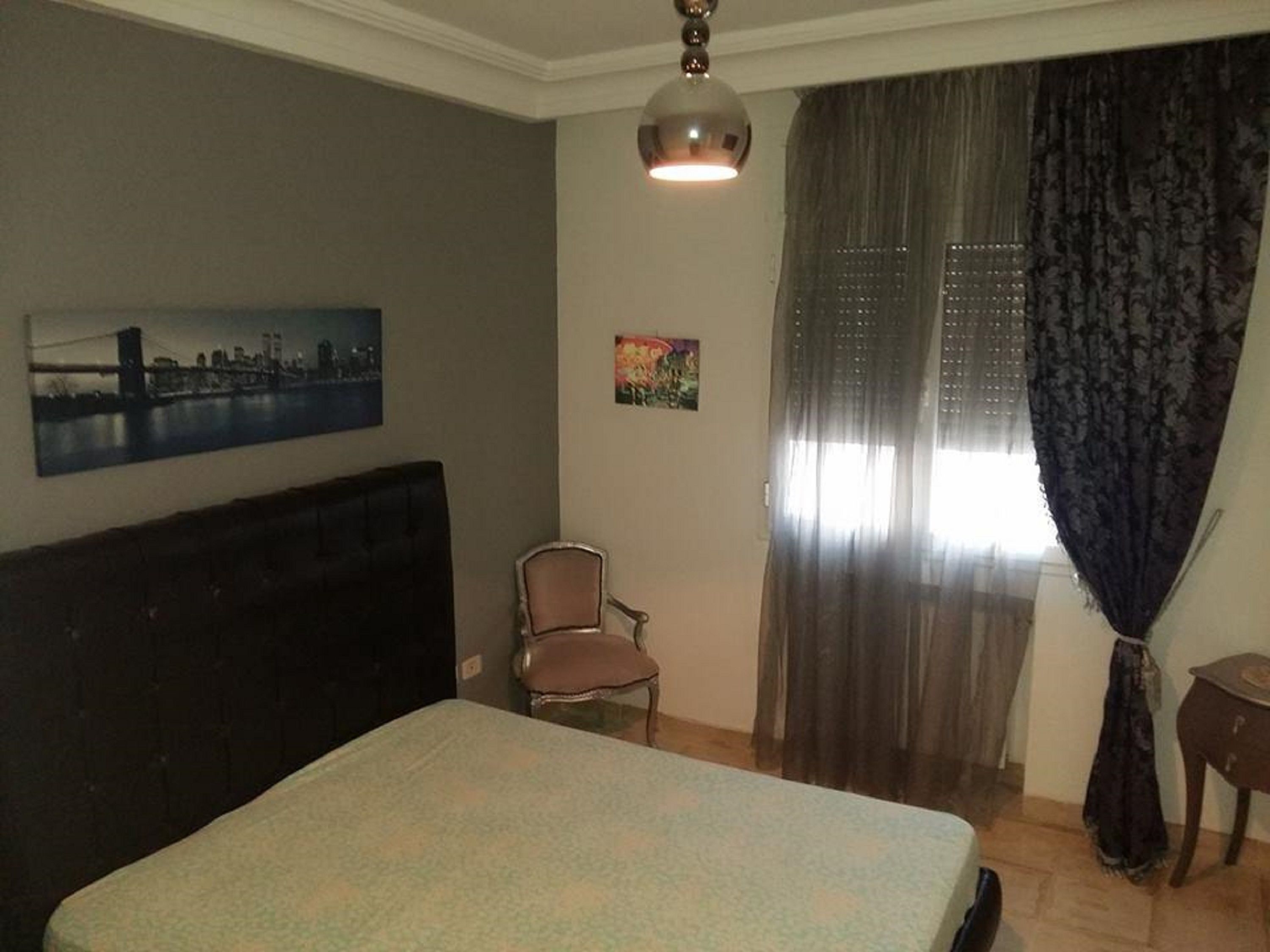 Une chambre à coucher avec un lit, une chaise, un bureau, un tableau et une fenêtre avec des rideaux.
