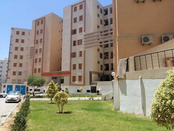 A multi-story residential building with a flat facade, multiple balconies, and a small garden area with young trees. - Benzineb Immo Menzeh (Ariana)