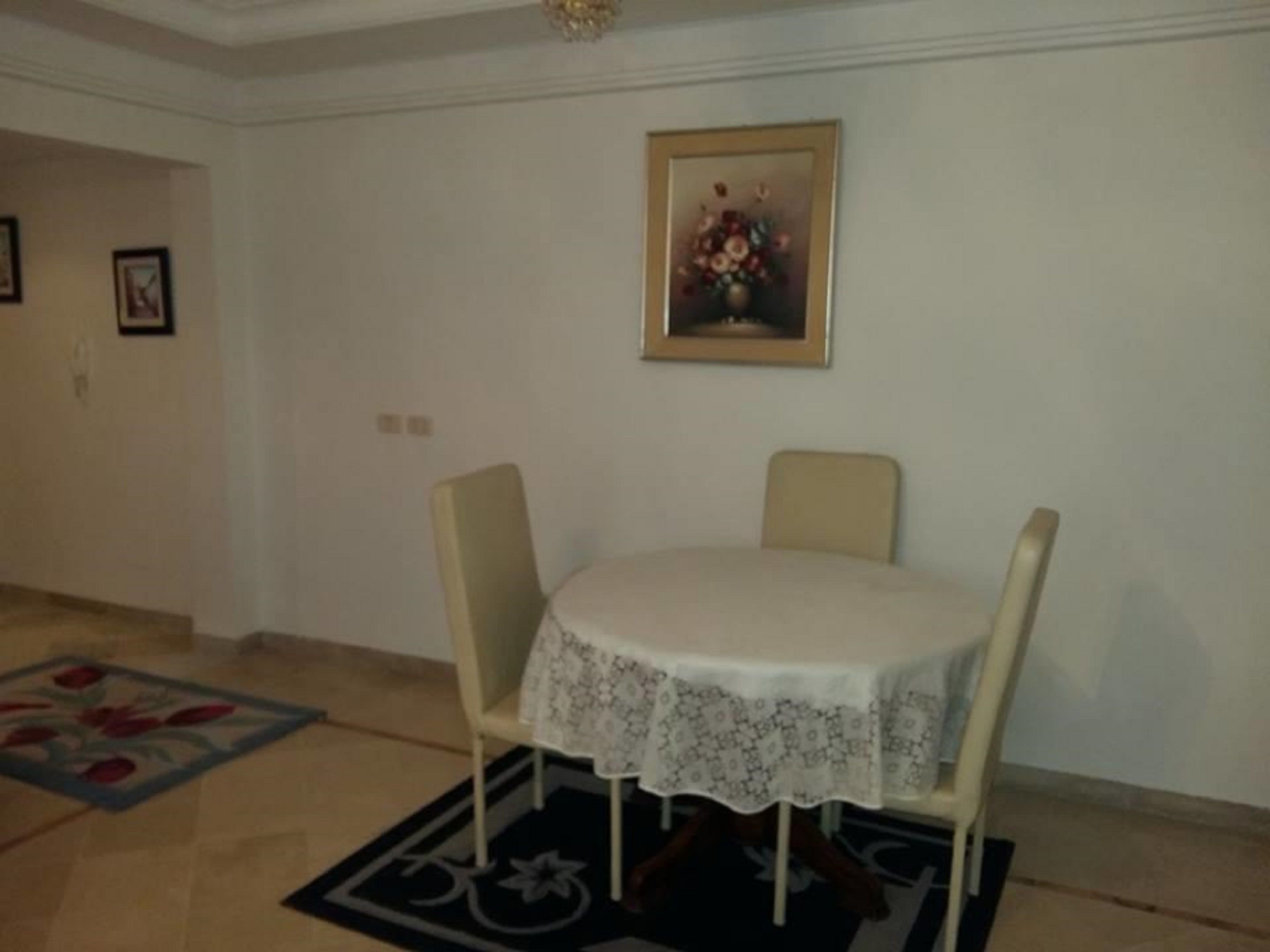 Un coin repas avec une table ronde recouverte d’une nappe en dentelle, flanquée de deux chaises. Un tableau floral encadré est accroché au mur au-dessus de la table.