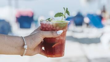 A hand holding a drink with a mint garnish, set against a blurred background of a beach with umbrellas.