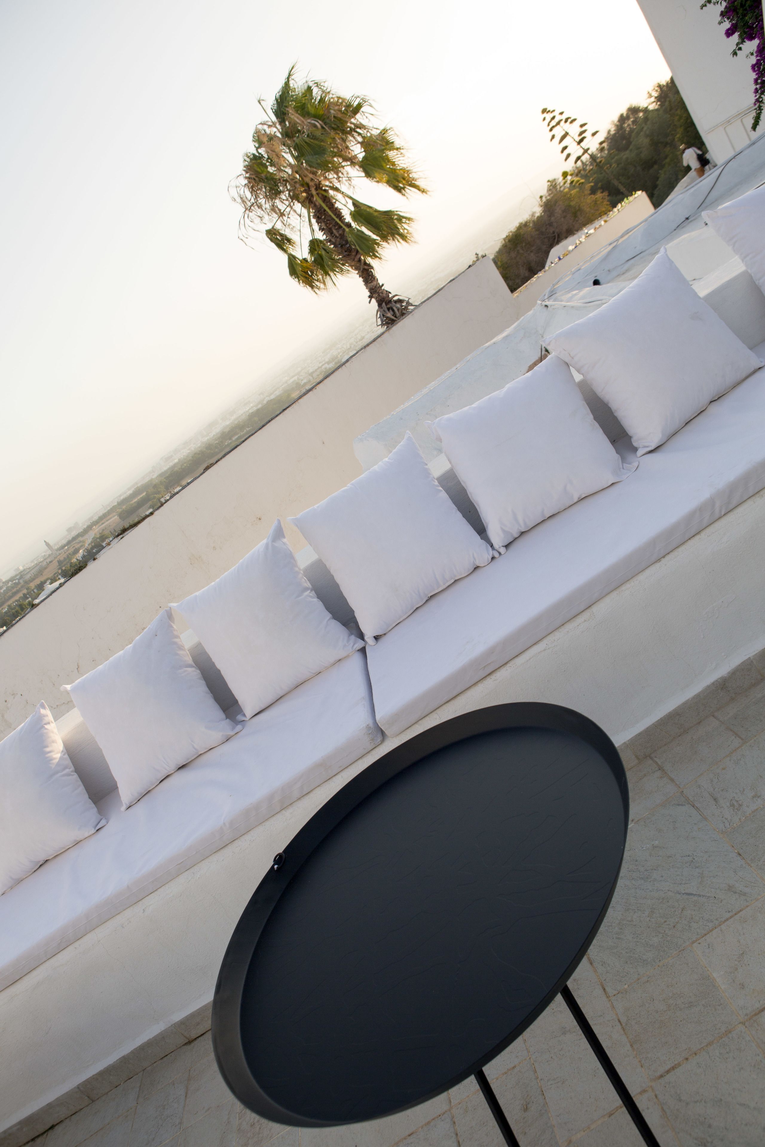 Une chaise longue avec des coussins blancs et une table noire, donnant sur une plage bordée de palmiers et sous un ciel dégagé.