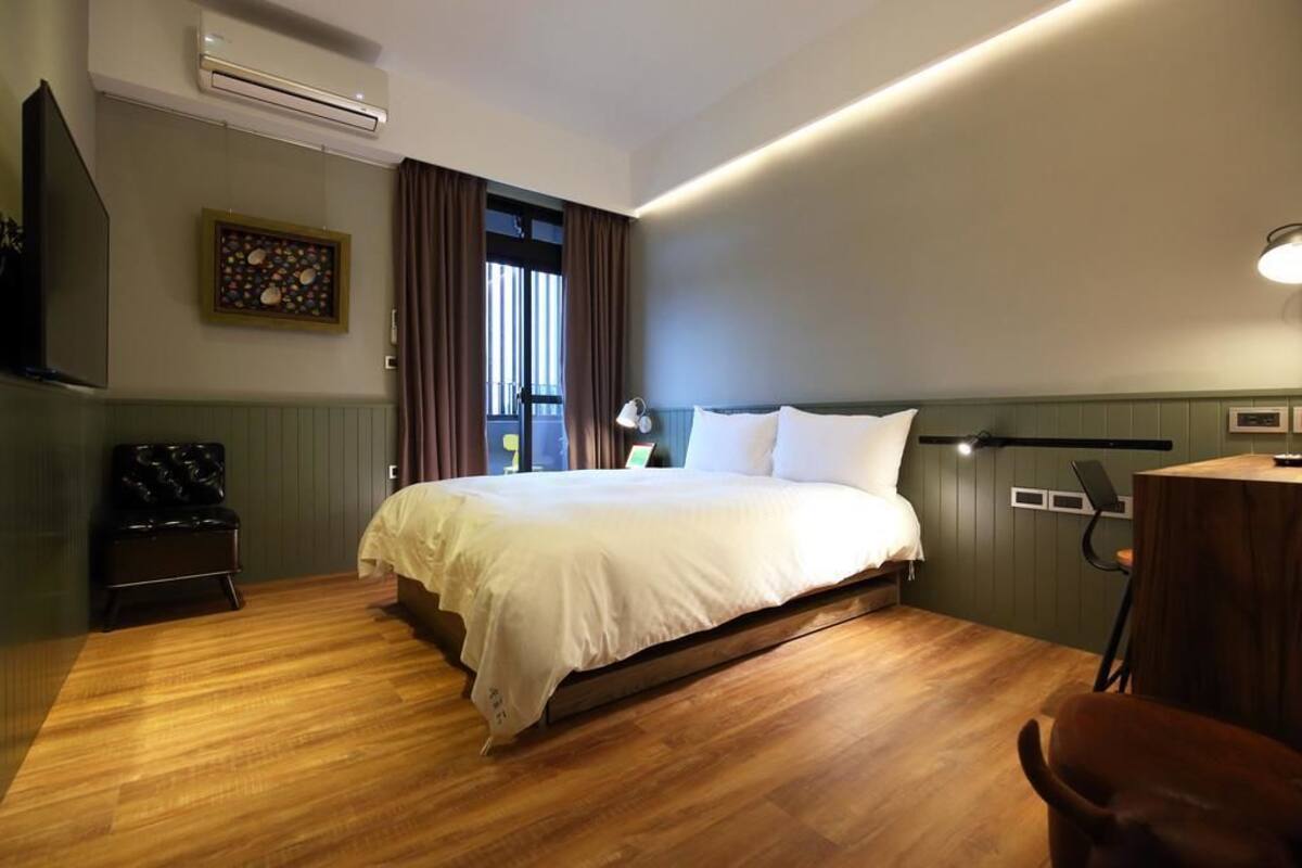 Una habitación de hotel moderna con piso de madera, una cama grande con ropa blanca, un escritorio con una silla y una ventana con cortinas.