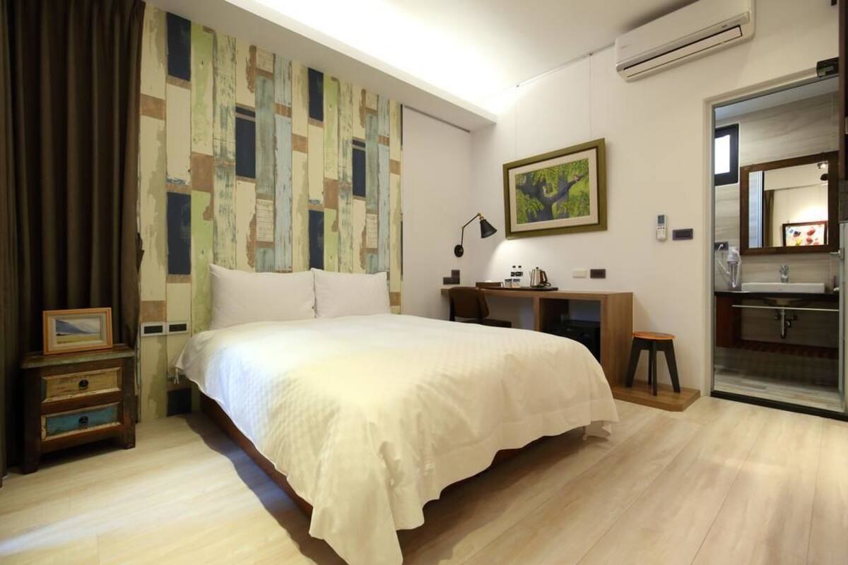 Un dormitorio con una cama grande, un escritorio, una silla, un taburete pequeño, una mesita de noche y un cuadro. Se ve un baño por una puerta abierta.
