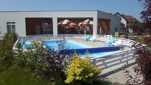 Piscina con cerco blanco, rodeada de vegetación y un edificio con zona de estar al aire libre.