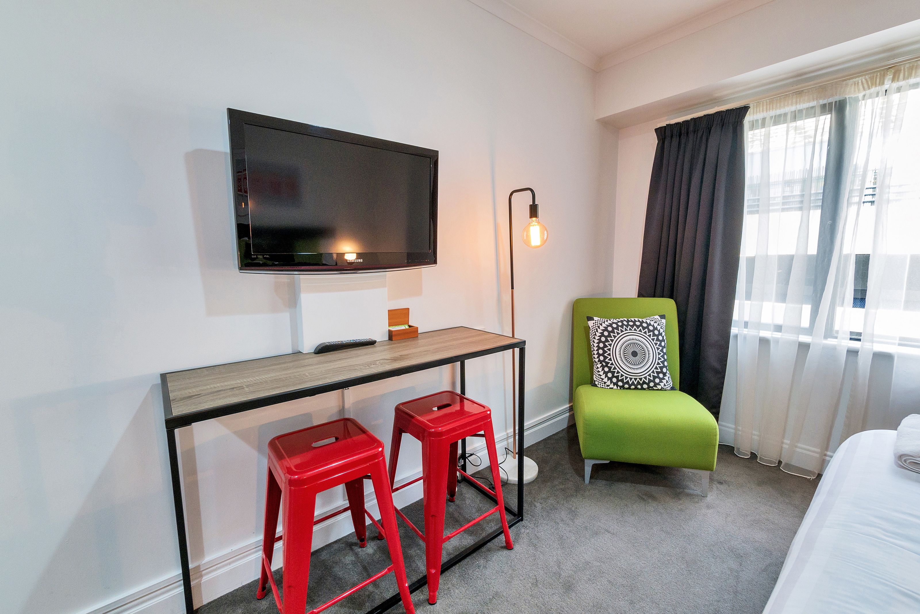 A hotel room with a TV mounted on the wall, a desk with two red stools, a green chair with a patterned pillow, and a bed with a white comforter.