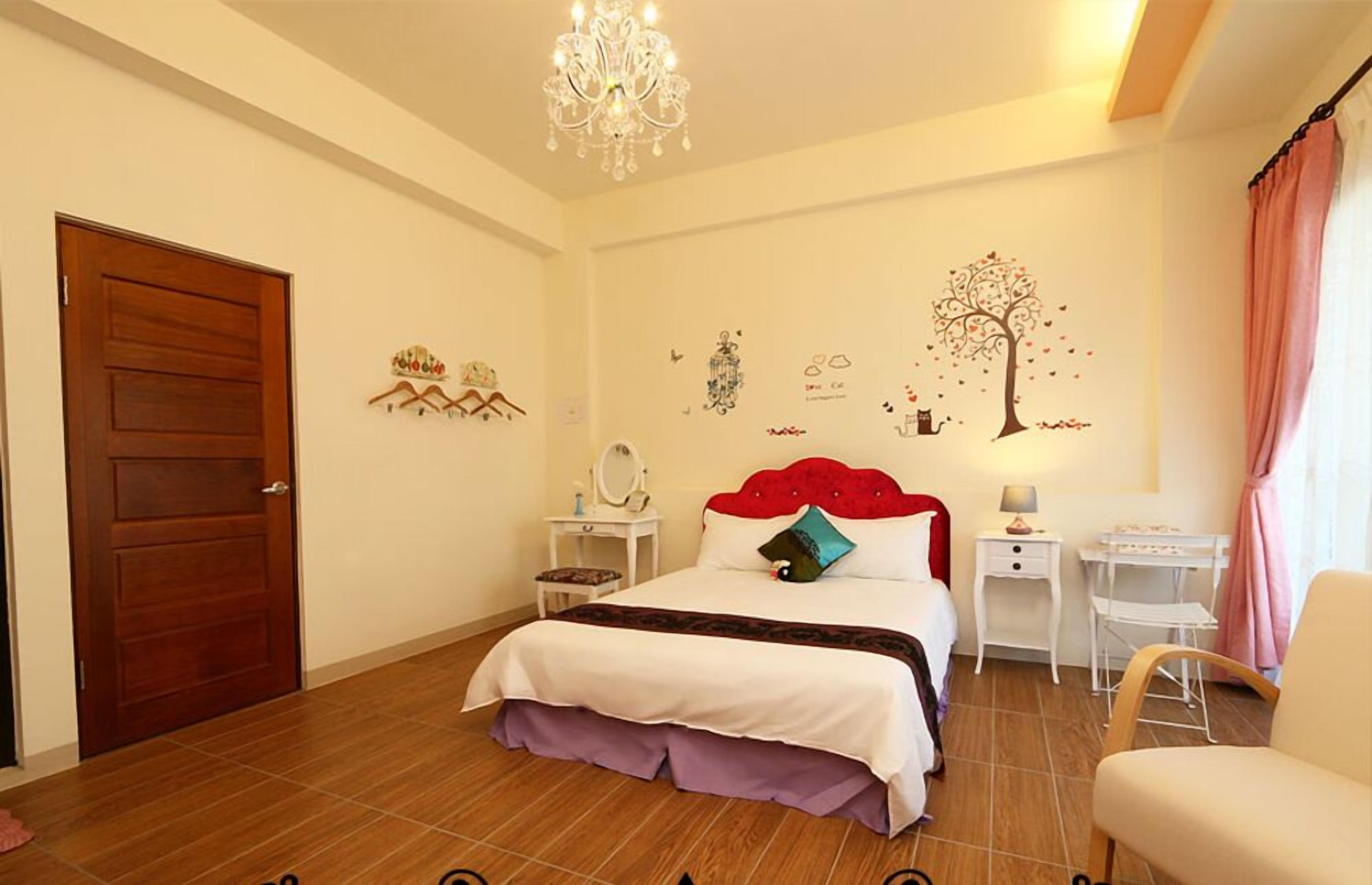 A bedroom with a wooden door, a bed with a red headboard, two bedside tables, a chandelier, and a wall with decorative stickers.