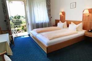 A hotel room with a large bed, two bedside tables with lamps, a small table with a chair, and a view of a balcony with flowers. - Walserhof (Wasserburg am Bodensee)