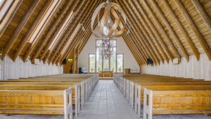 Interior de iglesia con bancales de madera dispuestos en filas, un alto techo de madera con una gran lámpara de araña, y un suelo de baldosas.