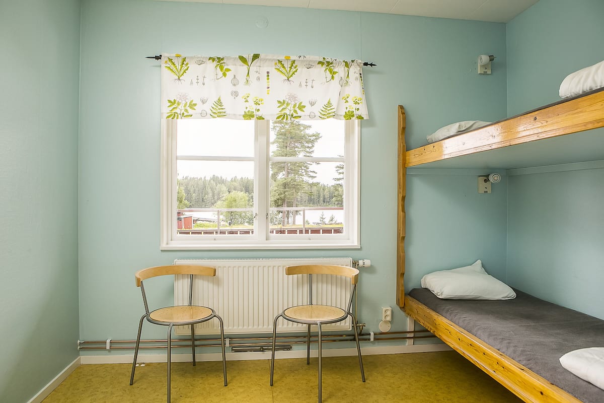 Un dormitorio con literas, un radiador, dos sillas y una ventana con cortina.