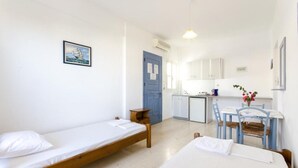 A small, clean room with two single beds, a wooden table with chairs, a small kitchenette, and a blue door. - Aris Apartments (Malia)