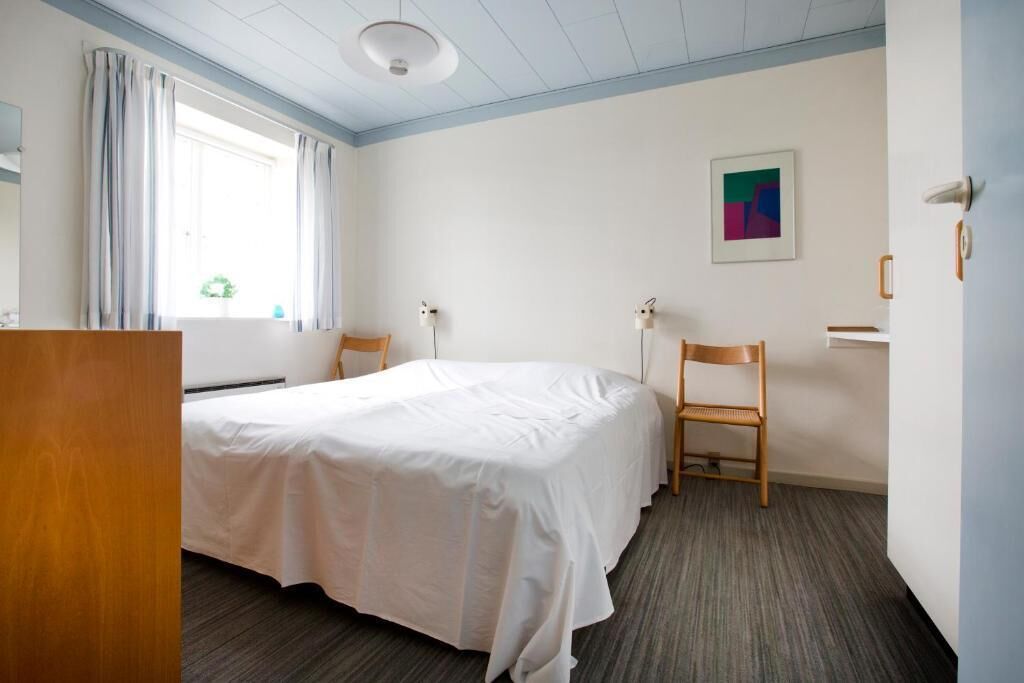 A hotel room with a bed, a desk, a chair, a desk lamp, a window with curtains, and a painting on the wall.