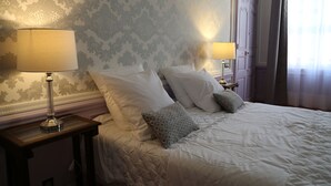 A neatly made bed with white linens and two pillows, a bedside table with a lamp, and a wall with patterned wallpaper. - Palais de Luppé (Arles)