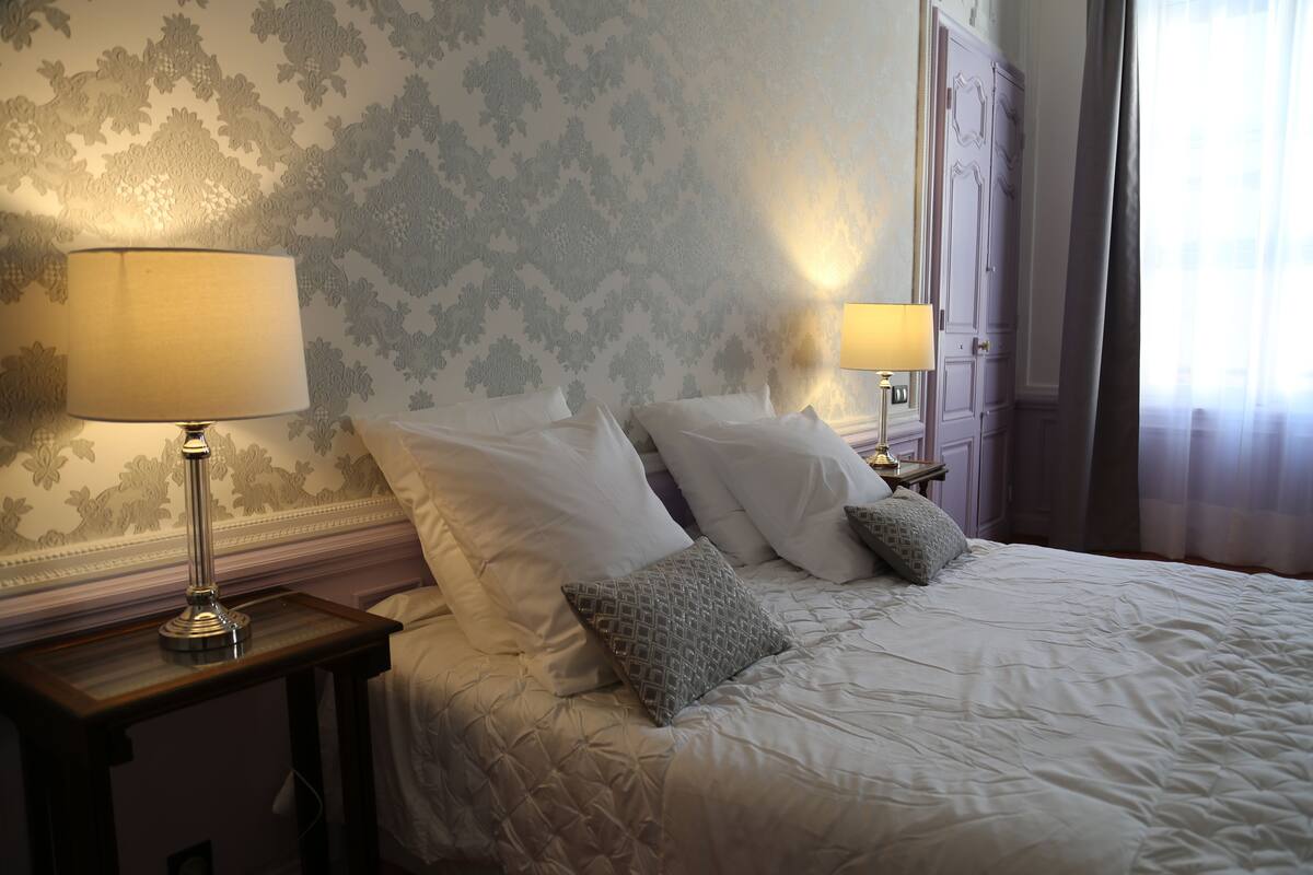 A neatly made bed with white linens and two pillows, a bedside table with a lamp, and a wall with patterned wallpaper.