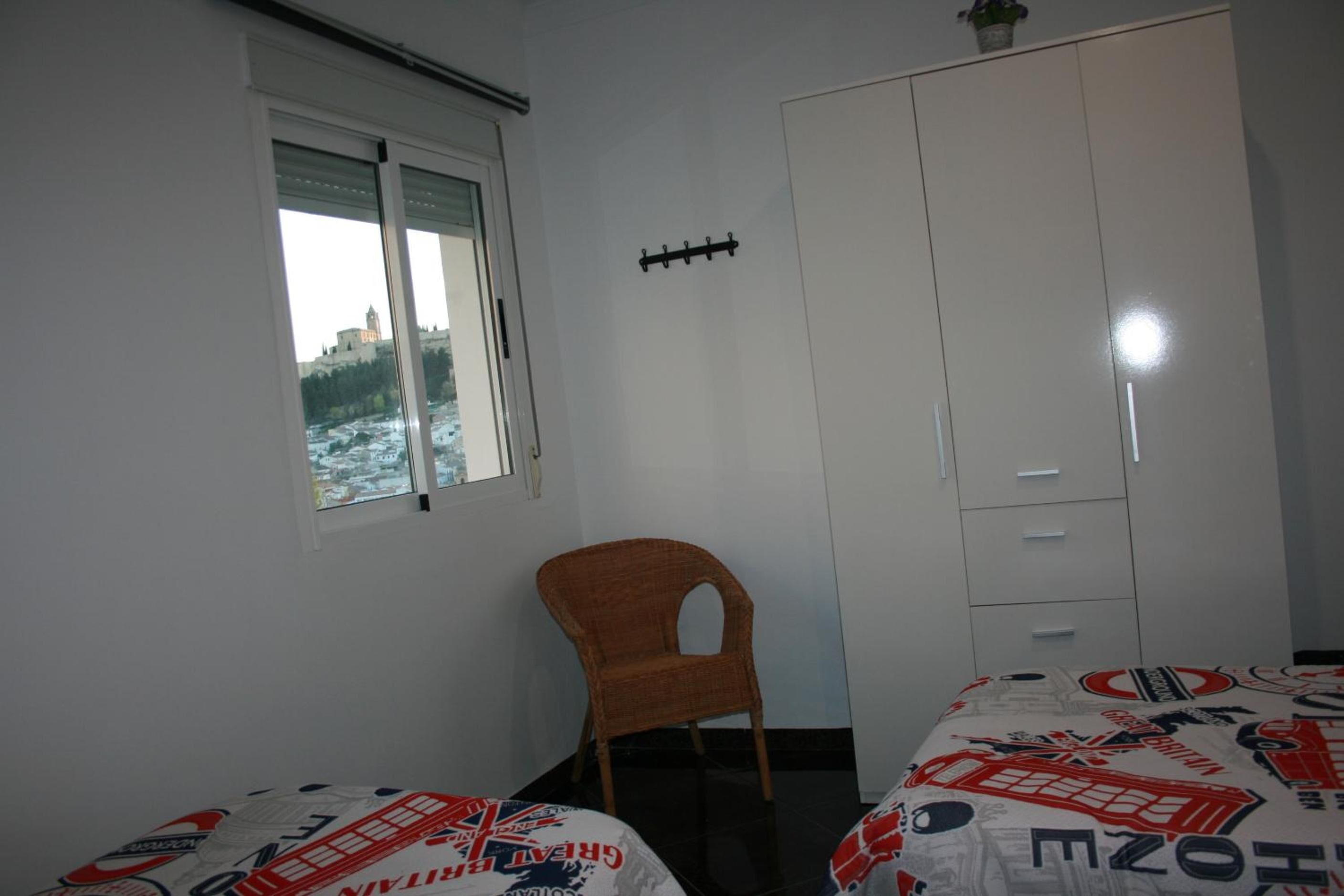A bedroom with two single beds, a wardrobe, a chair, and a window with a view of a building and trees.
