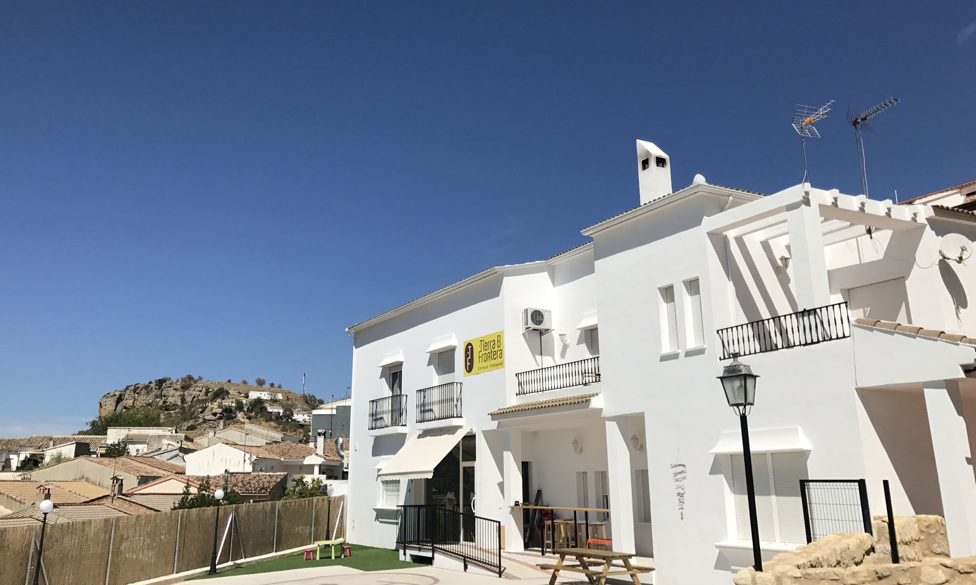Un edificio blanco con balcones, un letrero que dice "Tierra B Frontera" y una vista de una colina al fondo.