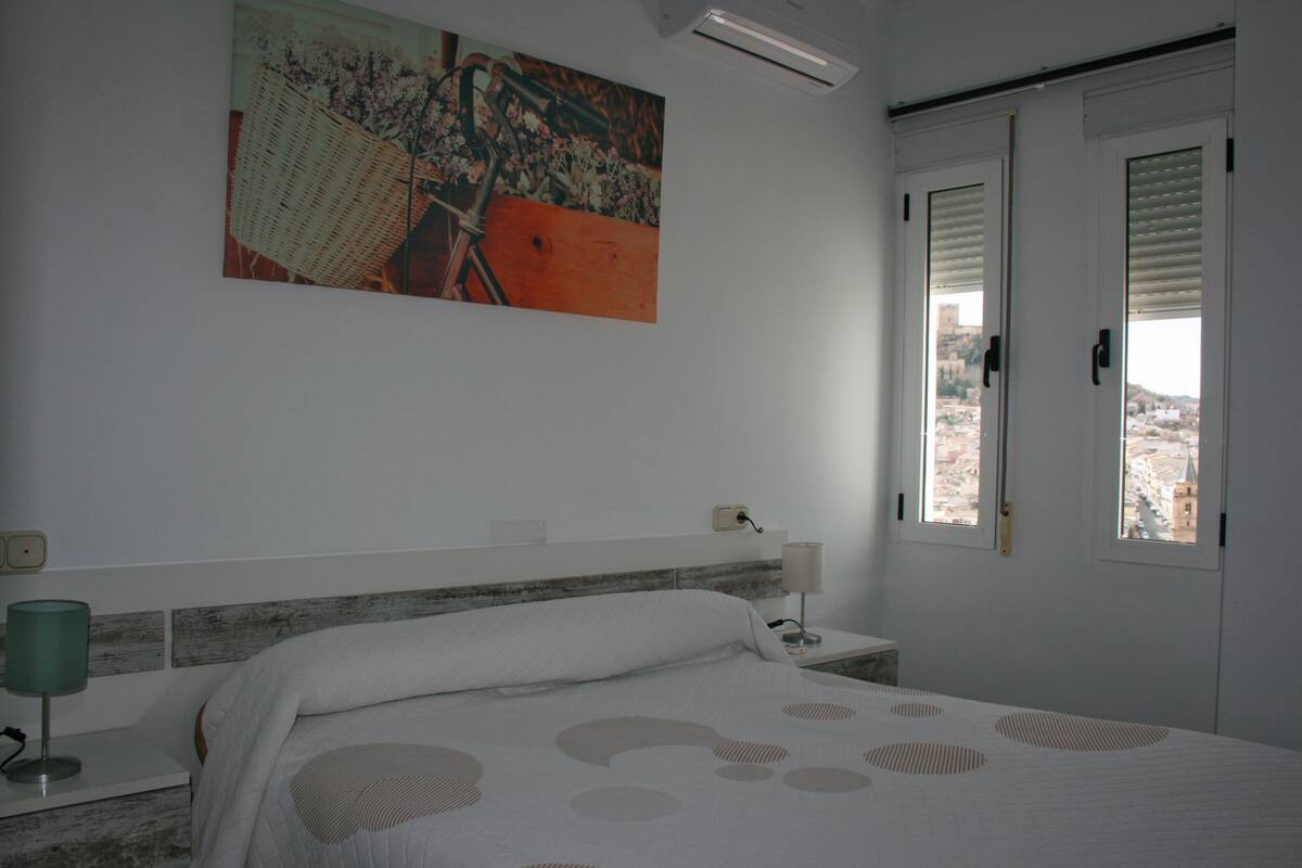 Une chambre à coucher comprenant un lit, une table de chevet, une lampe, un tableau et une fenêtre donnant sur un paysage urbain.