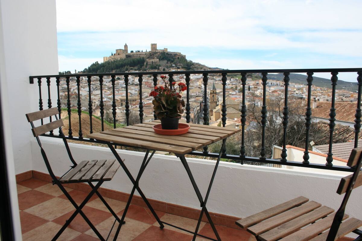 Un balcon avec une table et des chaises en bois, un pot de fleurs et une vue sur un château historique perché sur une colline.