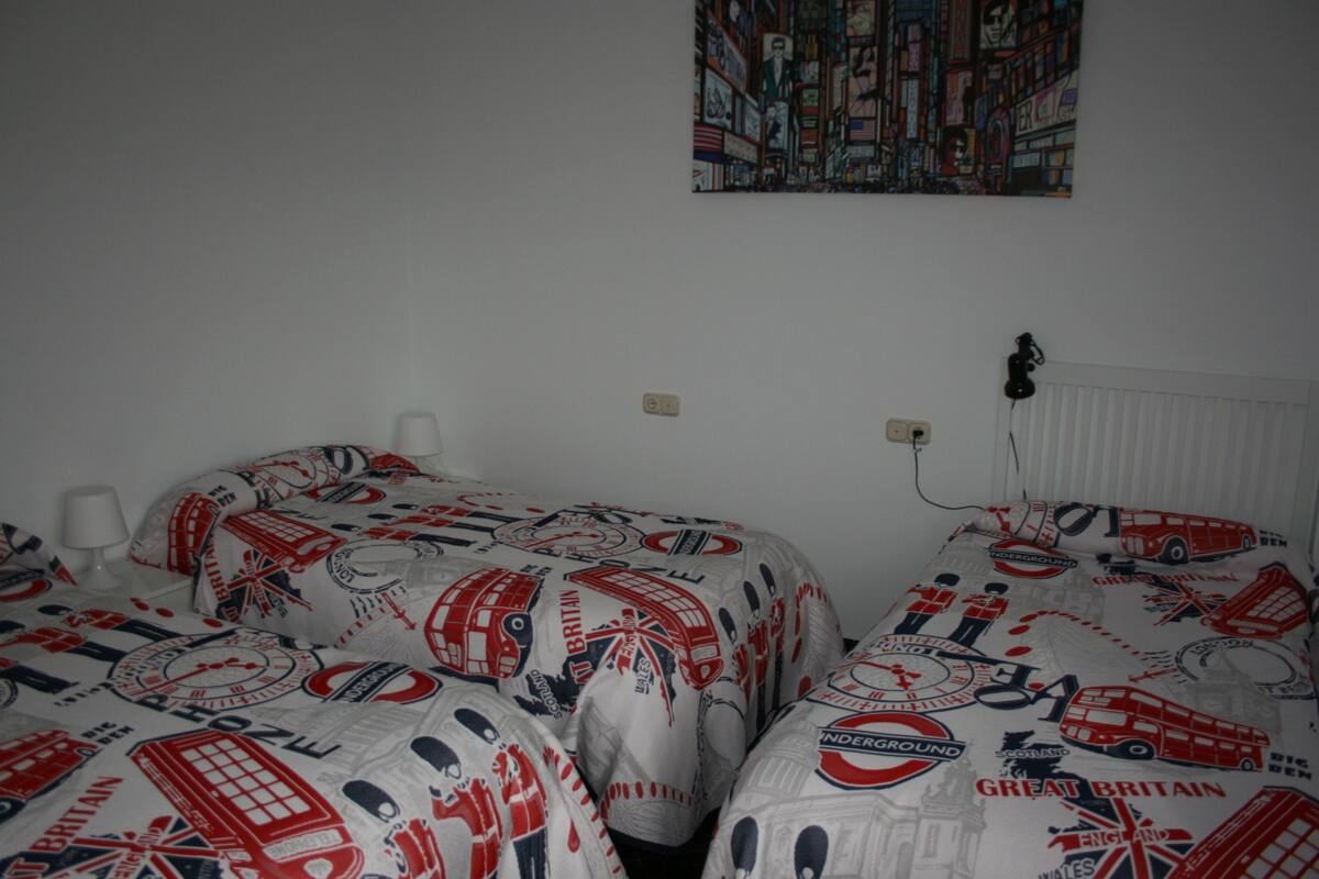 Une chambre avec deux lits, chacun recouvert d’une couette blanche ornée de motifs rouges et bleus sur le thème de la Grande-Bretagne. Un tableau est accroché au mur au-dessus des lits, et une prise de courant est visible sur le mur.