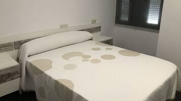 A neatly made bed with a patterned bedspread, a bedside table, and a window with blinds.