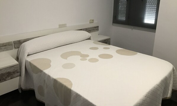 A neatly made bed with a patterned bedspread, a bedside table, and a window with blinds. - Mirador Tierra de Frontera (Alcala la Real)