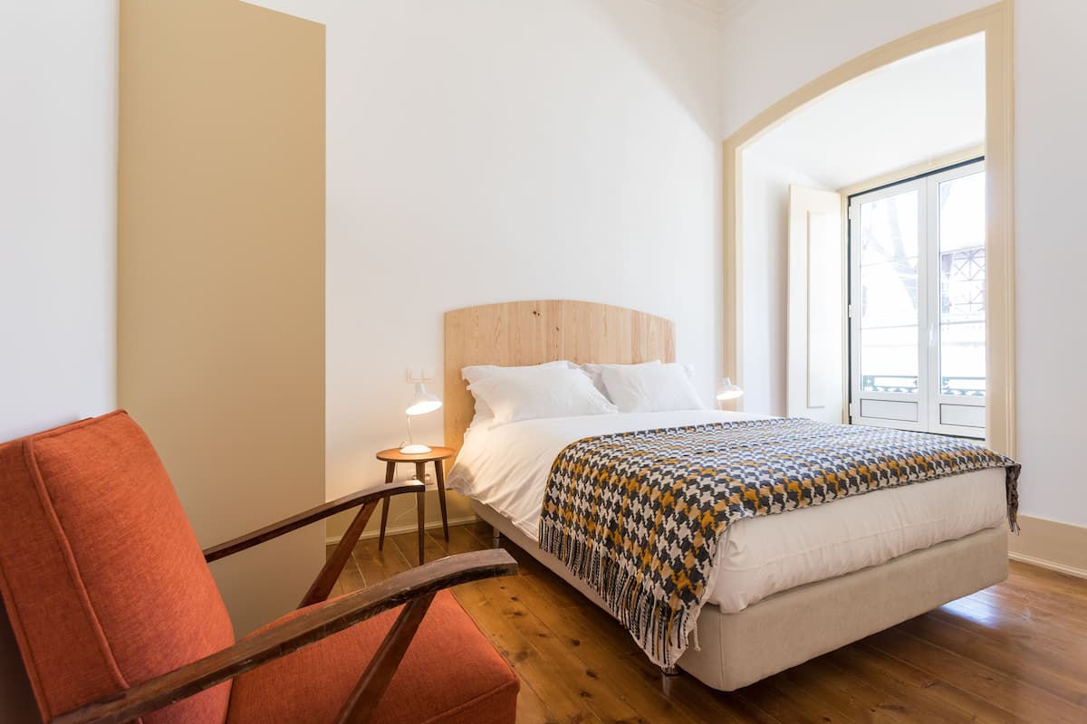 Un dormitorio con cabecera de madera, una cama con ropa de cama blanca, un plaid estampado, una silla de madera y una mesita de noche con una lámpara.