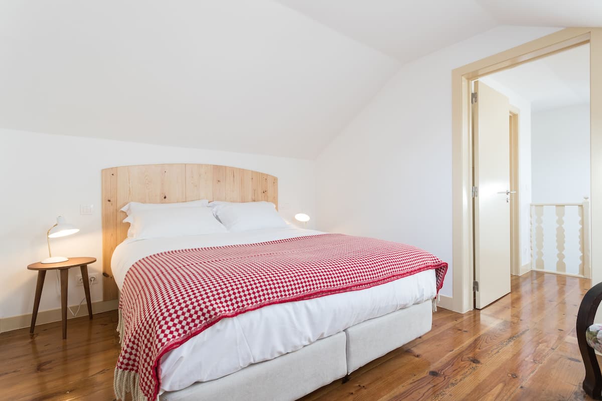 Un dormitorio con cabecera de madera, cama blanca con manta a cuadros rojos y blancos, mesita de noche con lámpara y suelo de madera.