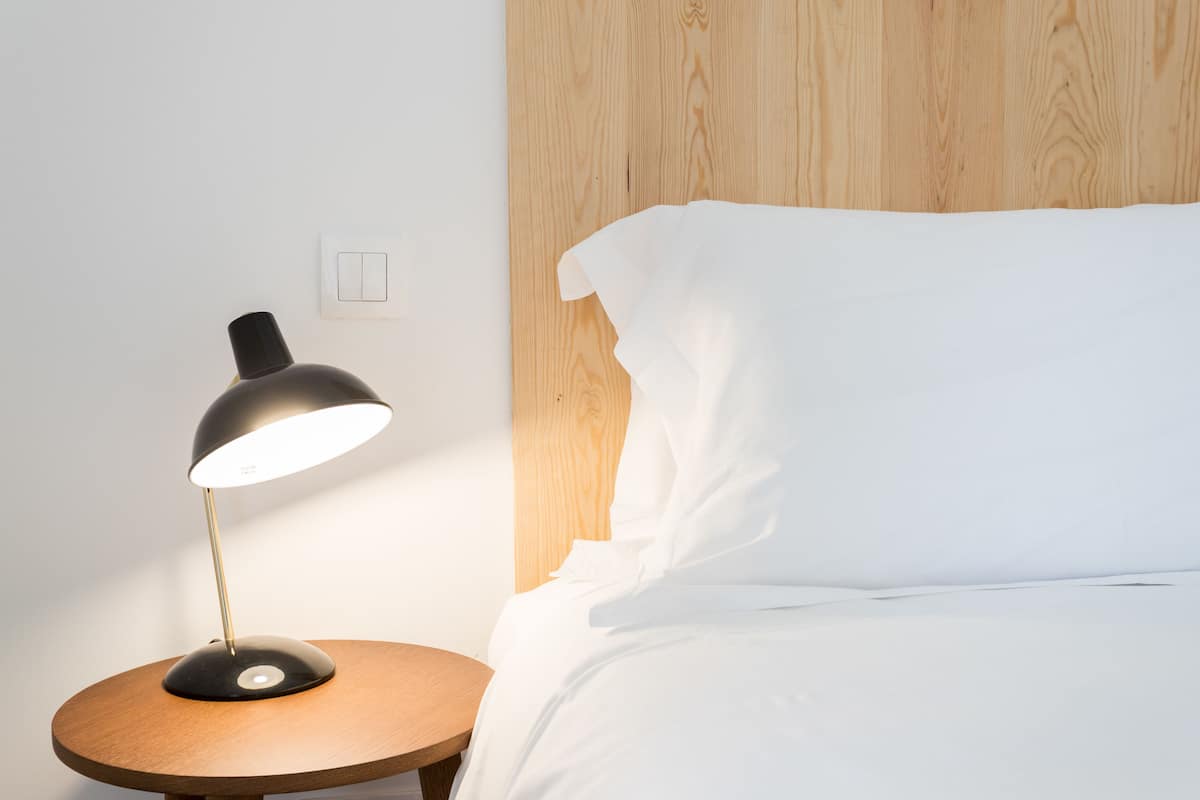 Una cama bien hecha con sábanas blancas, una mesita de noche redonda de madera con una lámpara negra, y un cabecero de madera.