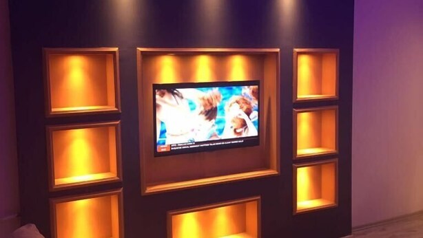 Una sala de estar moderna con un televisor de pantalla plana montado en una pared oscura, rodeada de estanterías iluminadas.