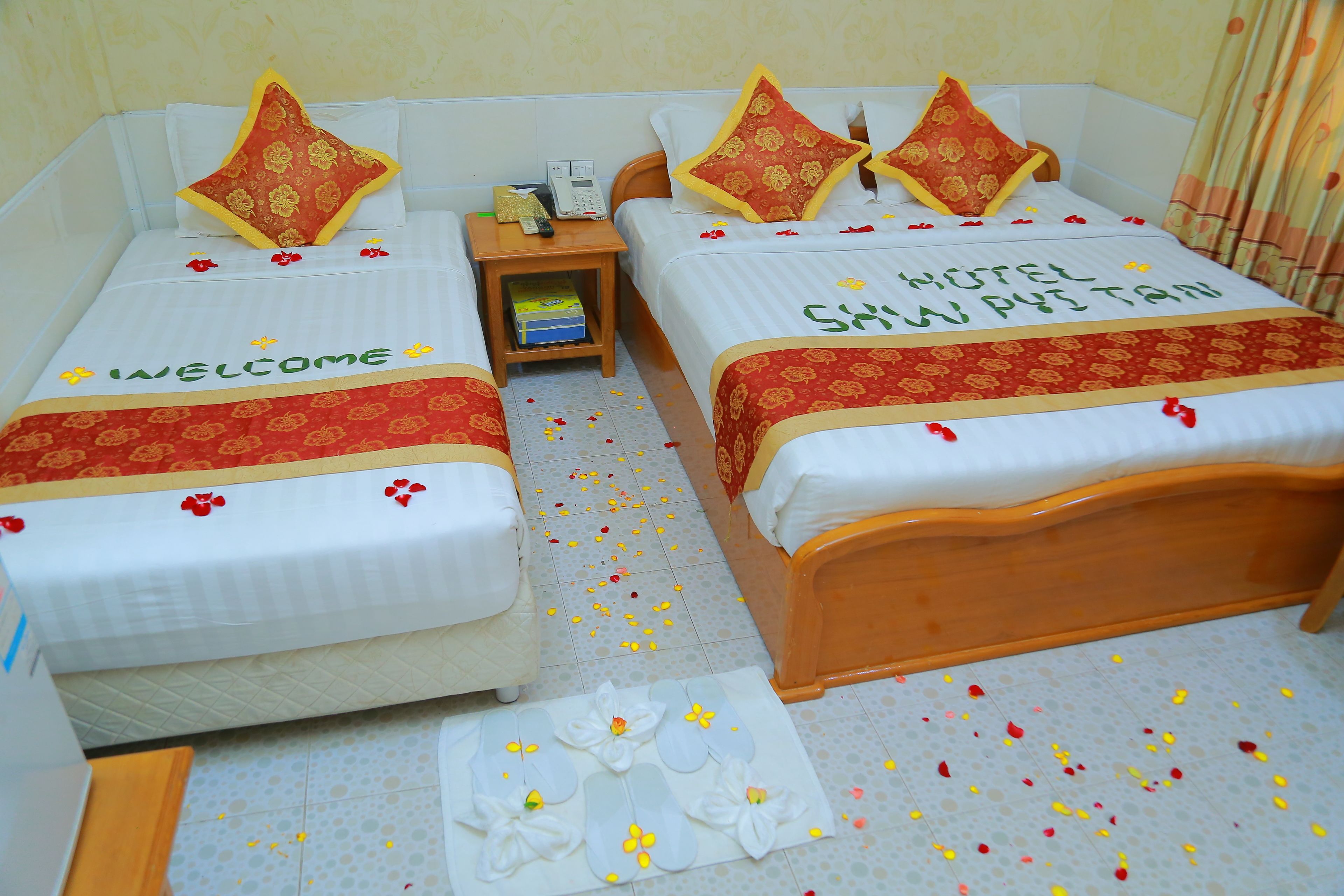 Two single beds with "WELCOME" and "HOTEL" text, a bedside table with a phone, and a small shelf with books.