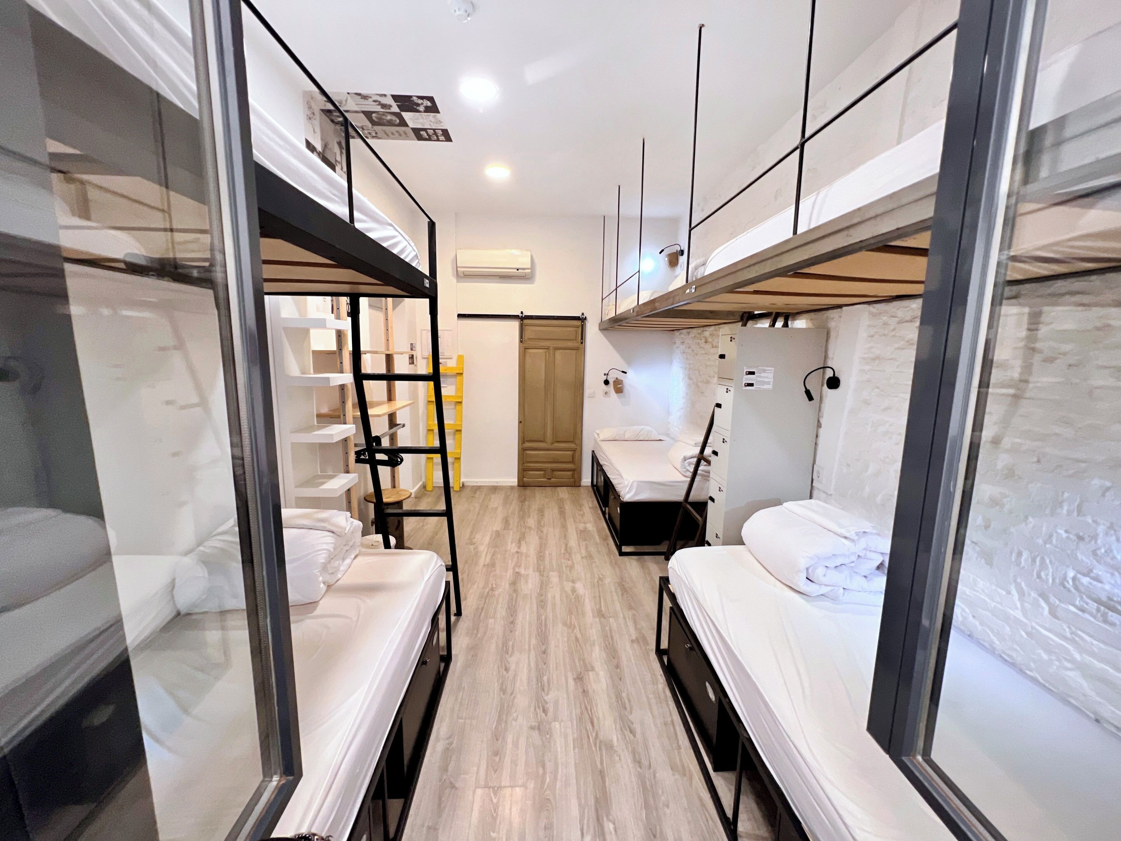Une chambre d’hôtel compacte avec des lits superposés, un plancher en bois et un petit bureau avec une chaise.