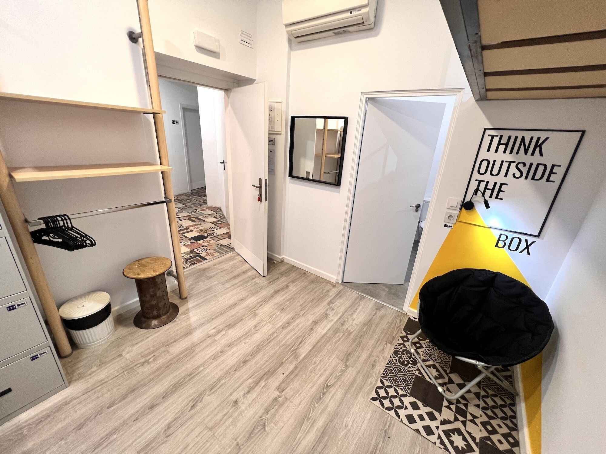 Habitación pequeña con suelo de madera, un taburete, estante con perchas, puerta con espejo y un cartel motivacional.