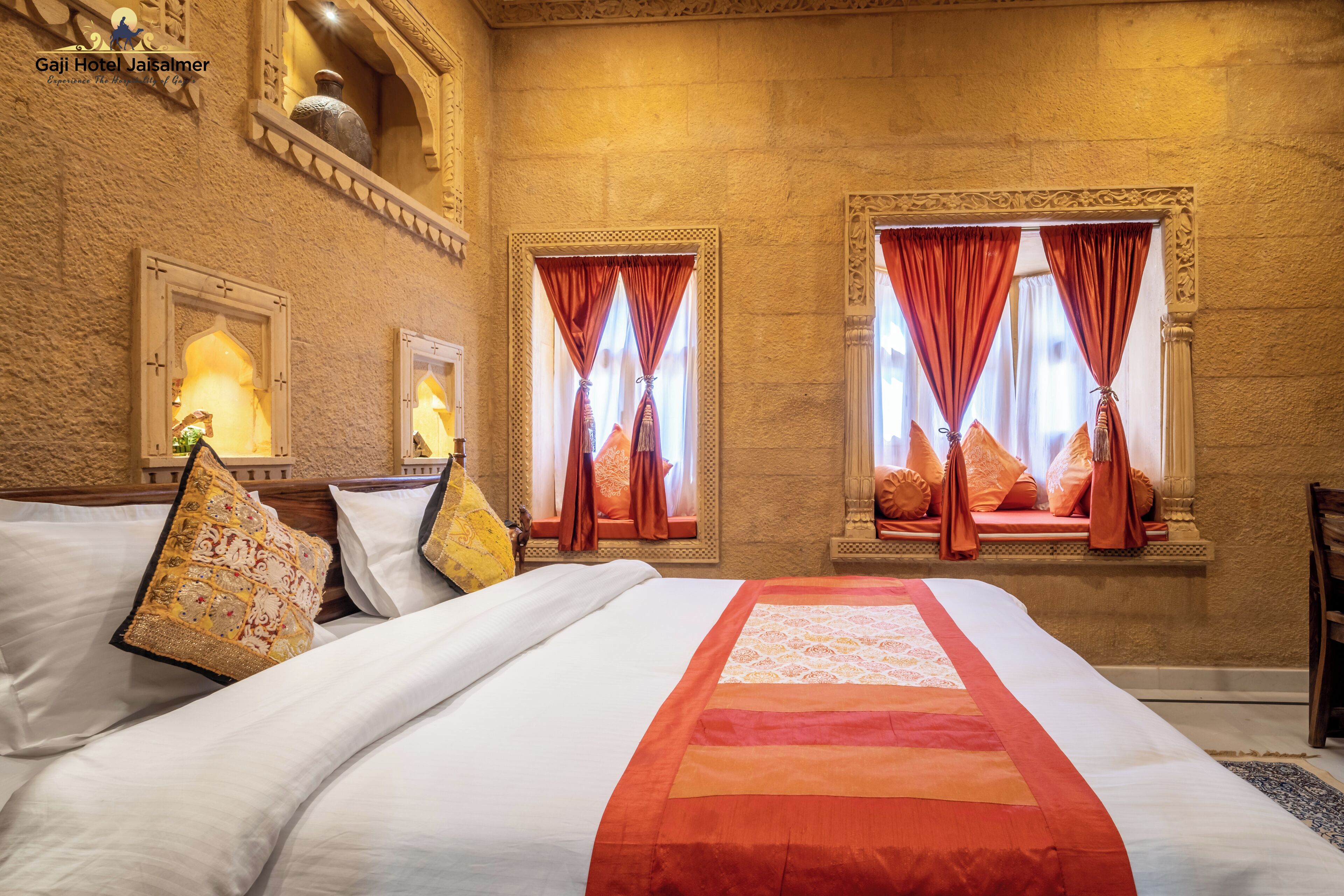 Habitación de hotel con una cama grande, almohadones decorativos y vistas de la ciudad a través de ventanas con cortinas rojas.