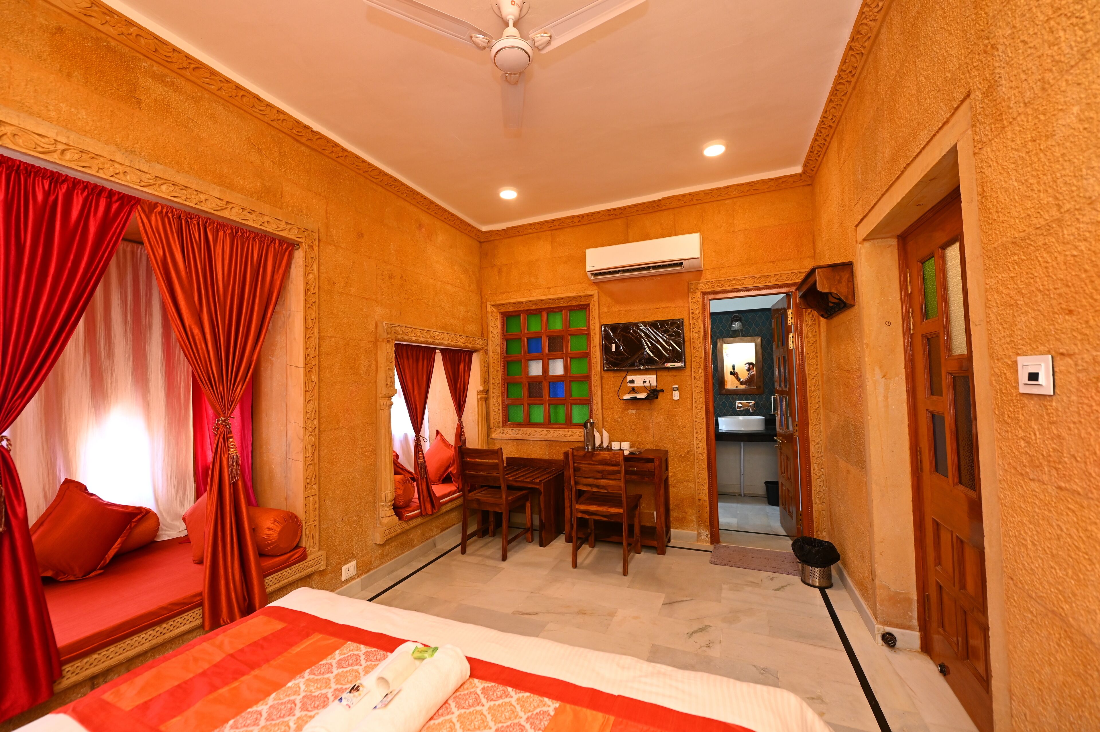 Quarto de hotel com cama, escrivaninha, cadeiras, janela com cortinas, ar condicionado e porta.