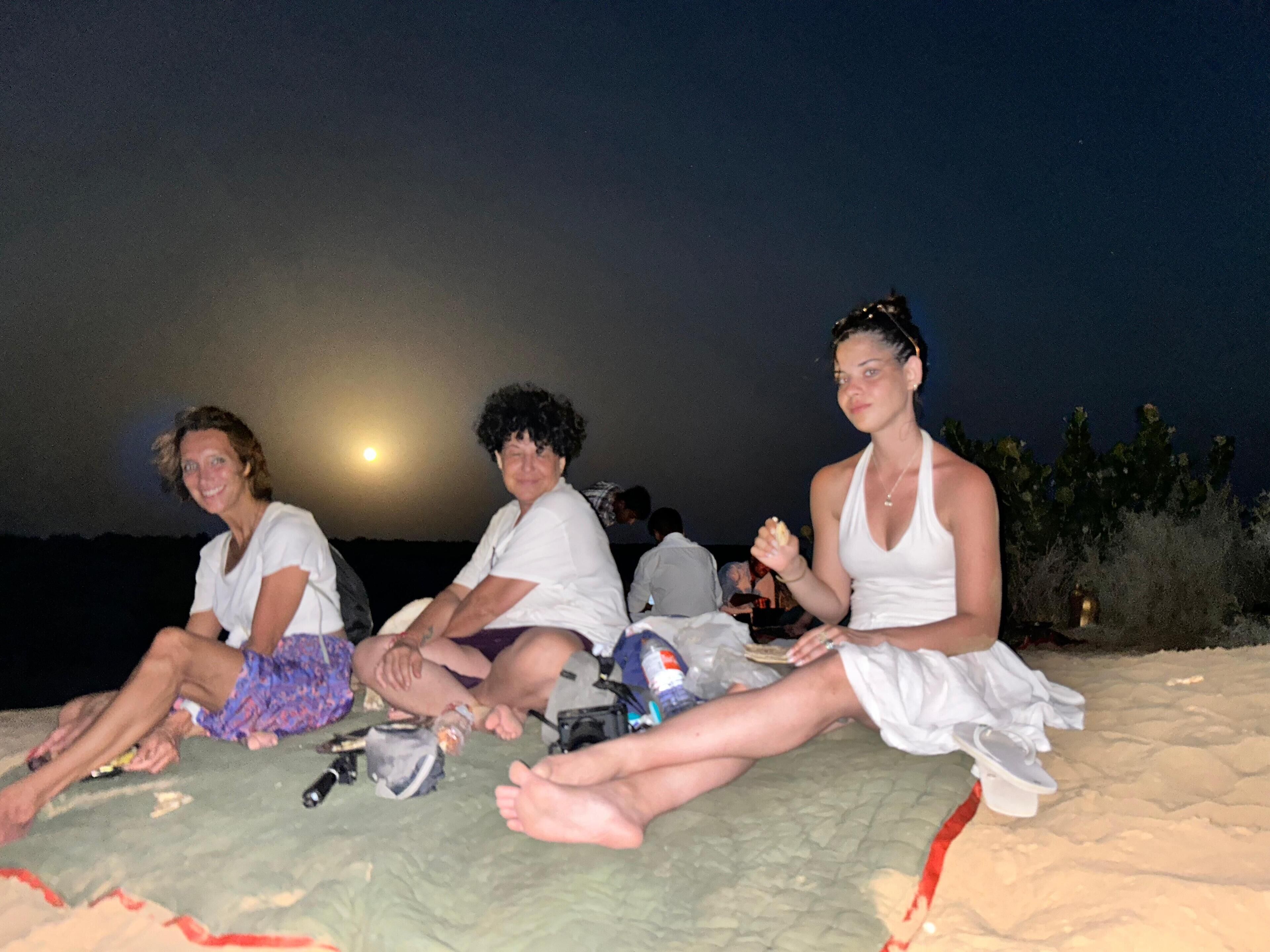 Drie mensen zitten op het strand in de nacht, met een maan op de achtergrond.