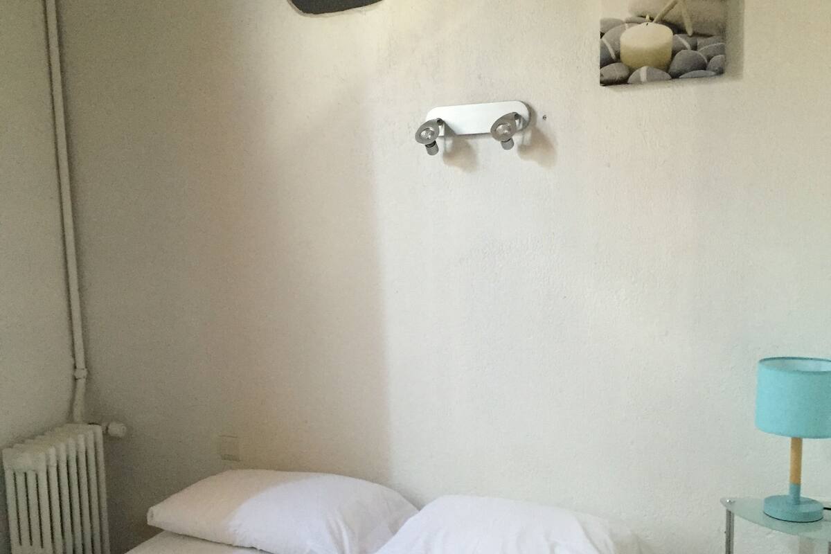 Una cama bien hecha con sábanas blancas, una mesita de noche con lámpara y un calentador de pared.