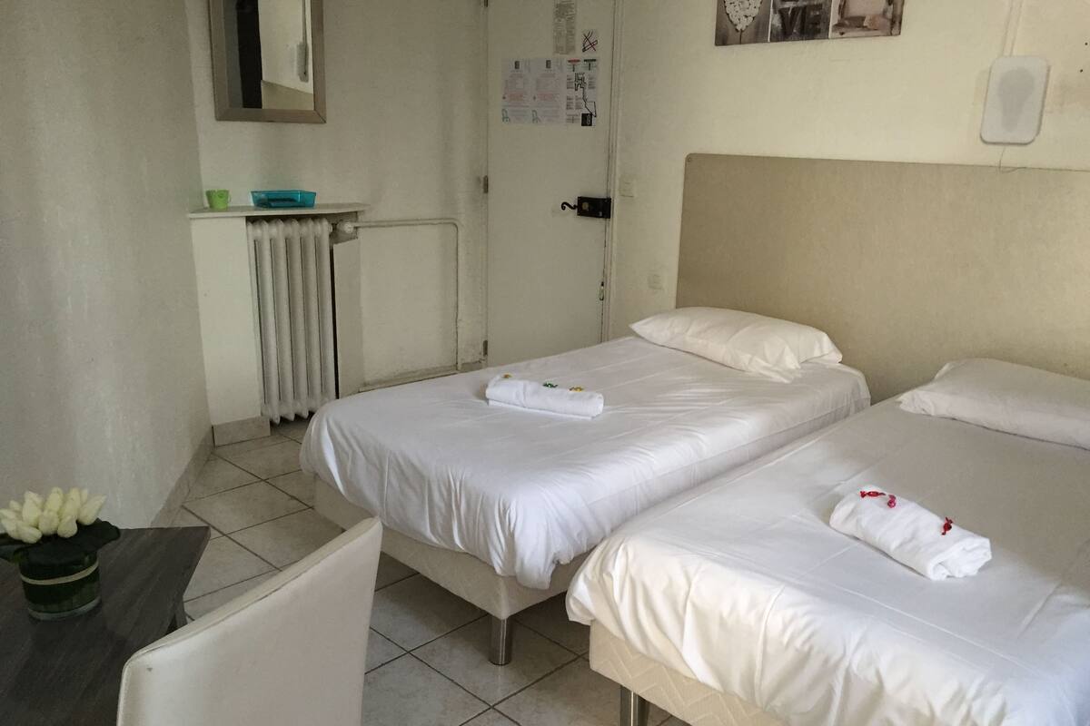 Habitación de hotel con dos camas, una mesita con una silla, un radiador y un espejo en la pared.