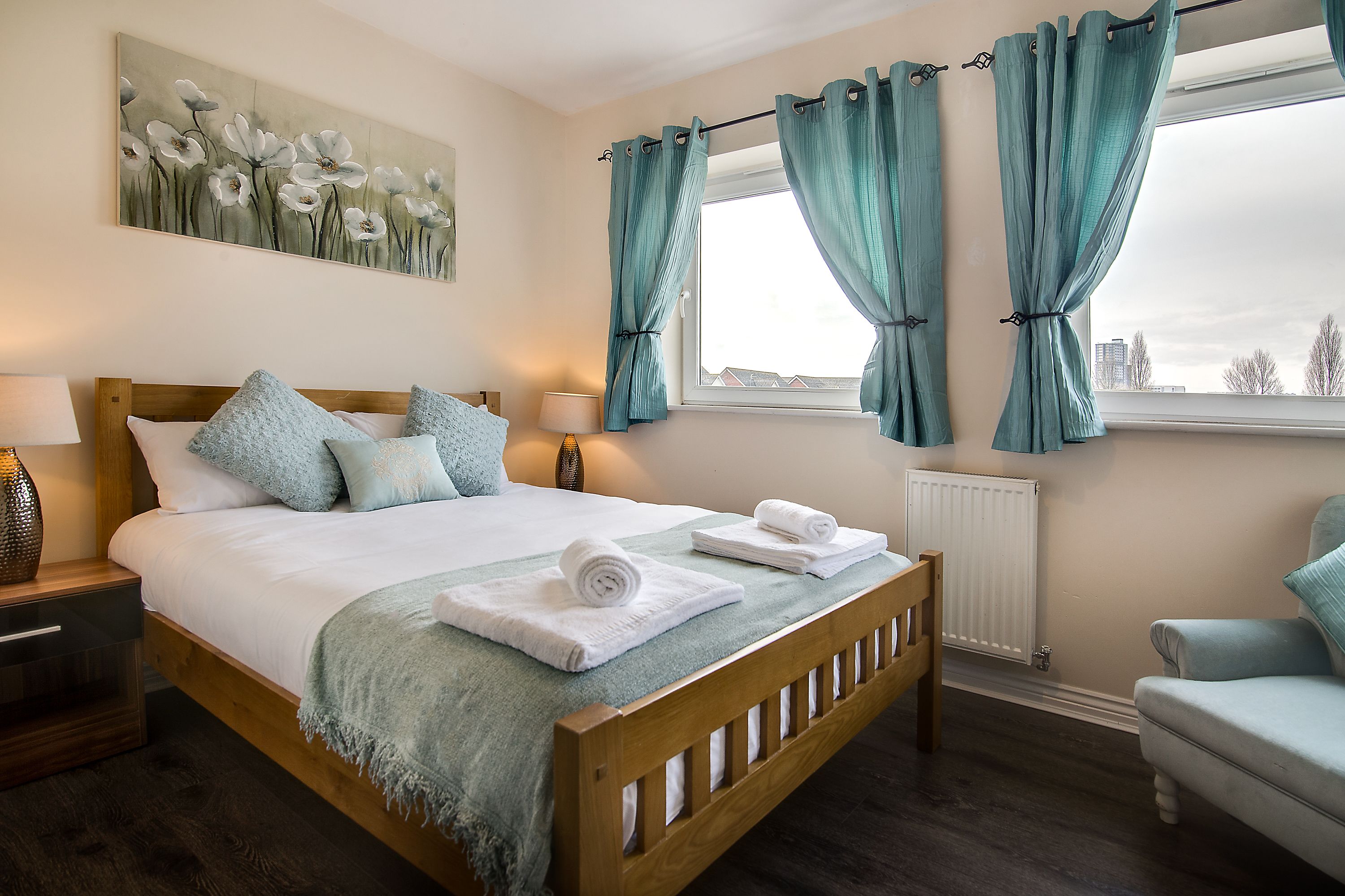 A neatly made bed with a white comforter and teal accents, a wooden headboard, and a view of the city through a window with teal curtains.