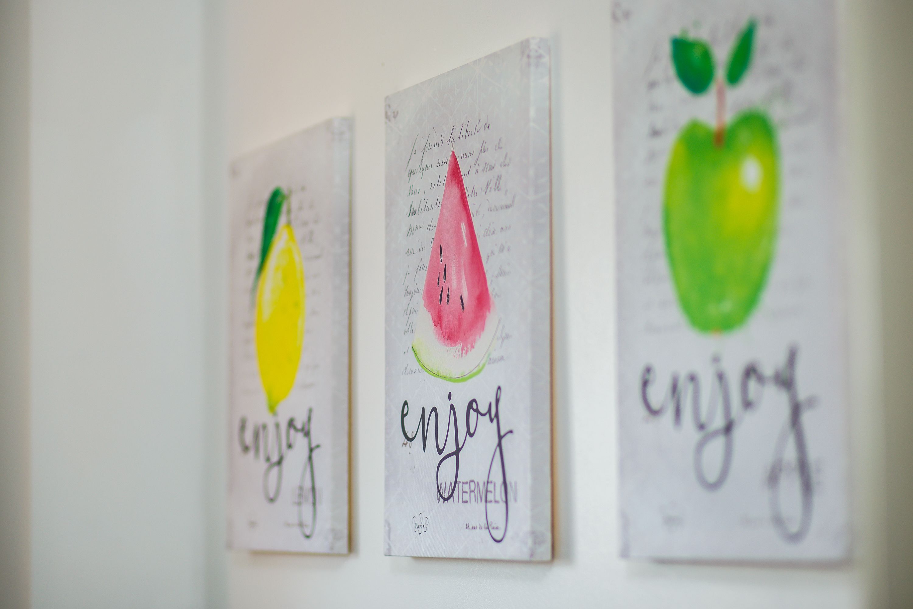 Three framed artworks with fruit illustrations and the word "enjoy" on a wall.