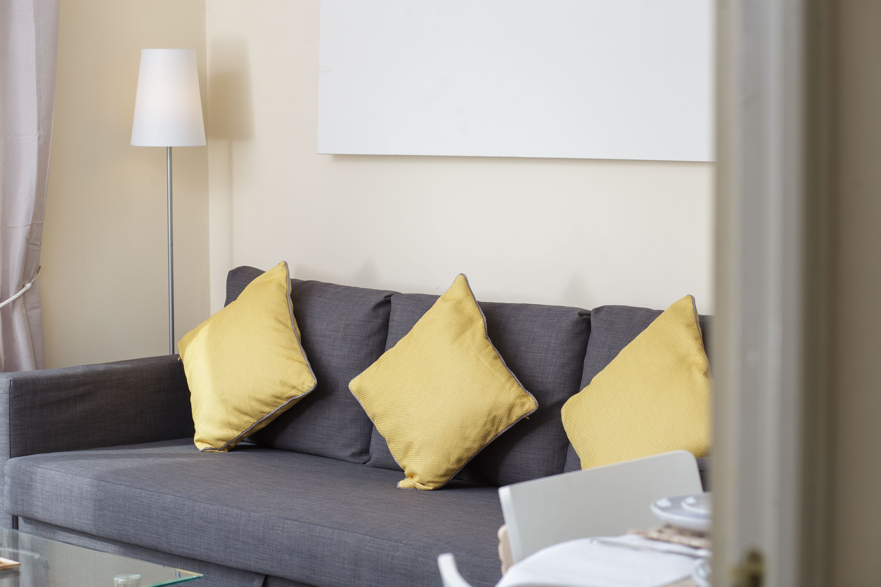 A grey sofa with three yellow cushions, a white lamp, and a laptop on a glass table.