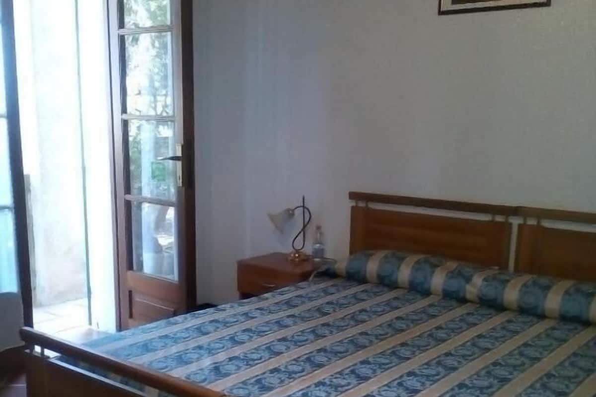 A bedroom with a wooden bed, a nightstand, a lamp, and a window with a view of greenery.