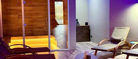 Quarto com piso de madeira, área de estar com cadeiras e uma mesa, e vista para área externa através de uma porta de vidro.