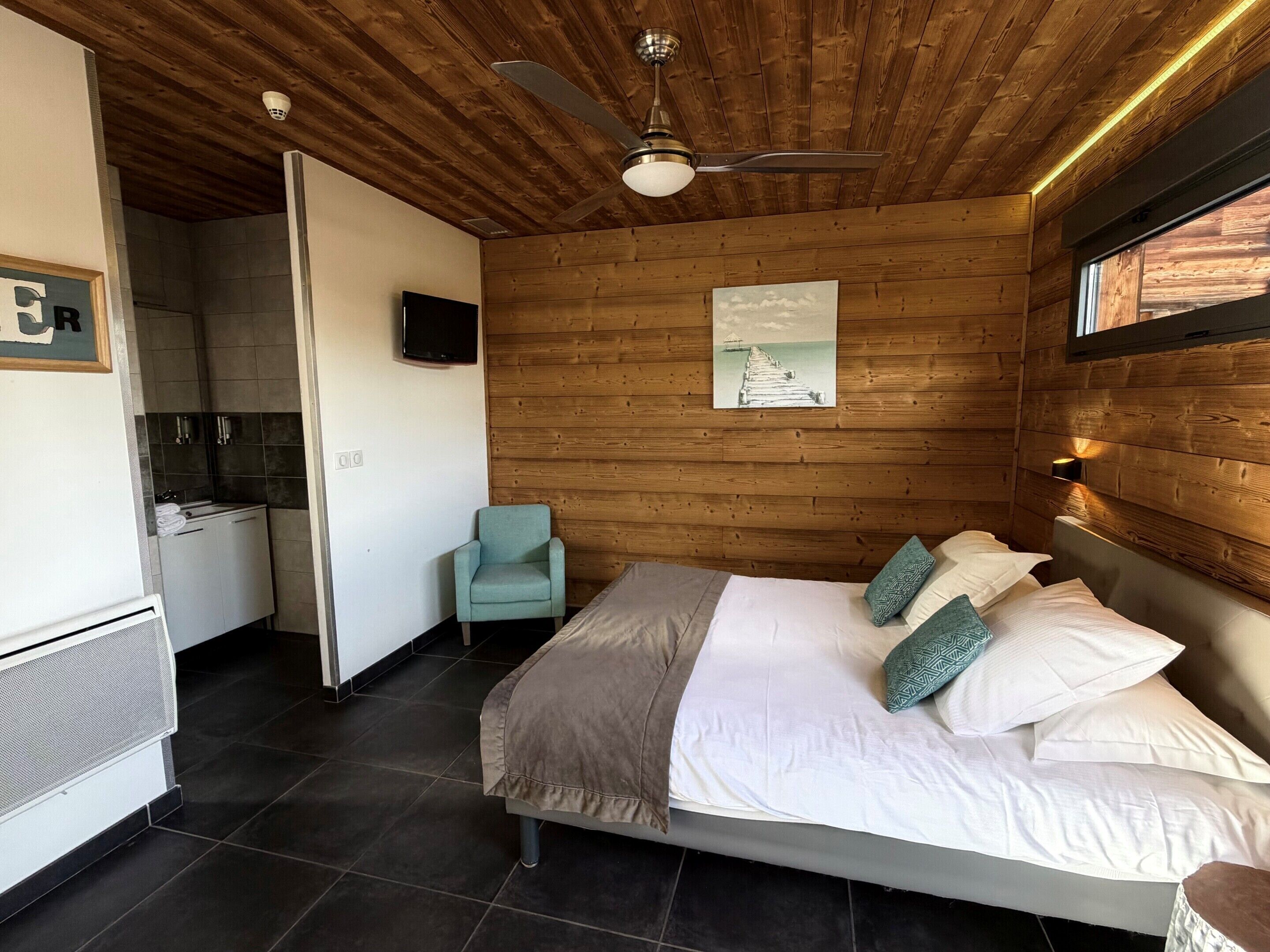 Un dormitorio con techo de madera, una cama con sábanas blancas, un sillón azul turquesa, un cuadro de un muelle y un televisor en la pared.